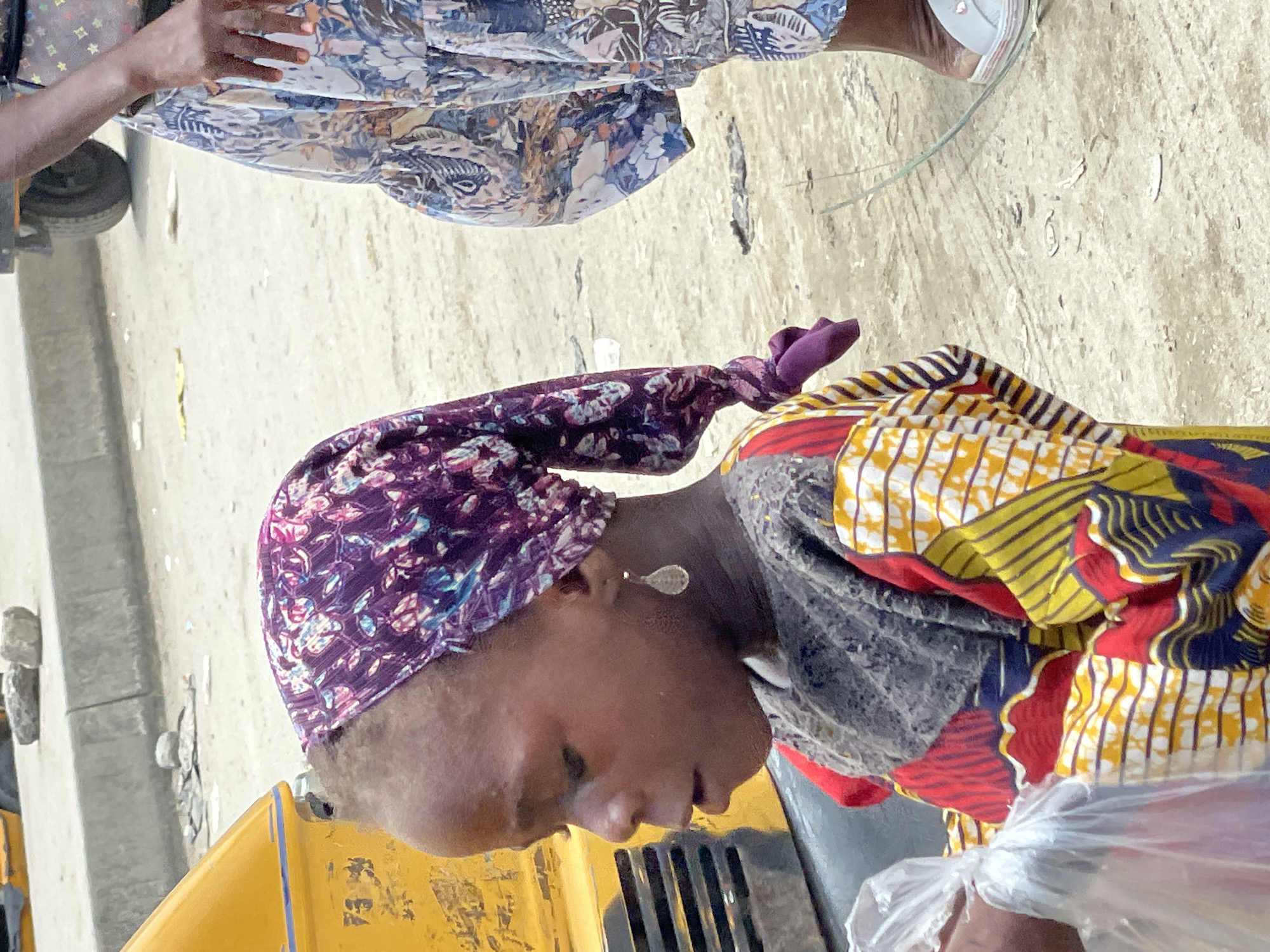 A woman is seen from an elevated perspective, wearing a purple headscarf with floral patterns and a patterned yellow, red, and navy top. She is positioned next to a yellow vehicle, possibly a commercial tricycle or auto-rickshaw, typical of the transport found in Lekki, Nigeria. Another person, out of focus, is present in the background with a floral patterned skirt. The ground is a pale concrete surface. A plastic bag is visible in the foreground, and a vehicle wheel can be seen in the background.