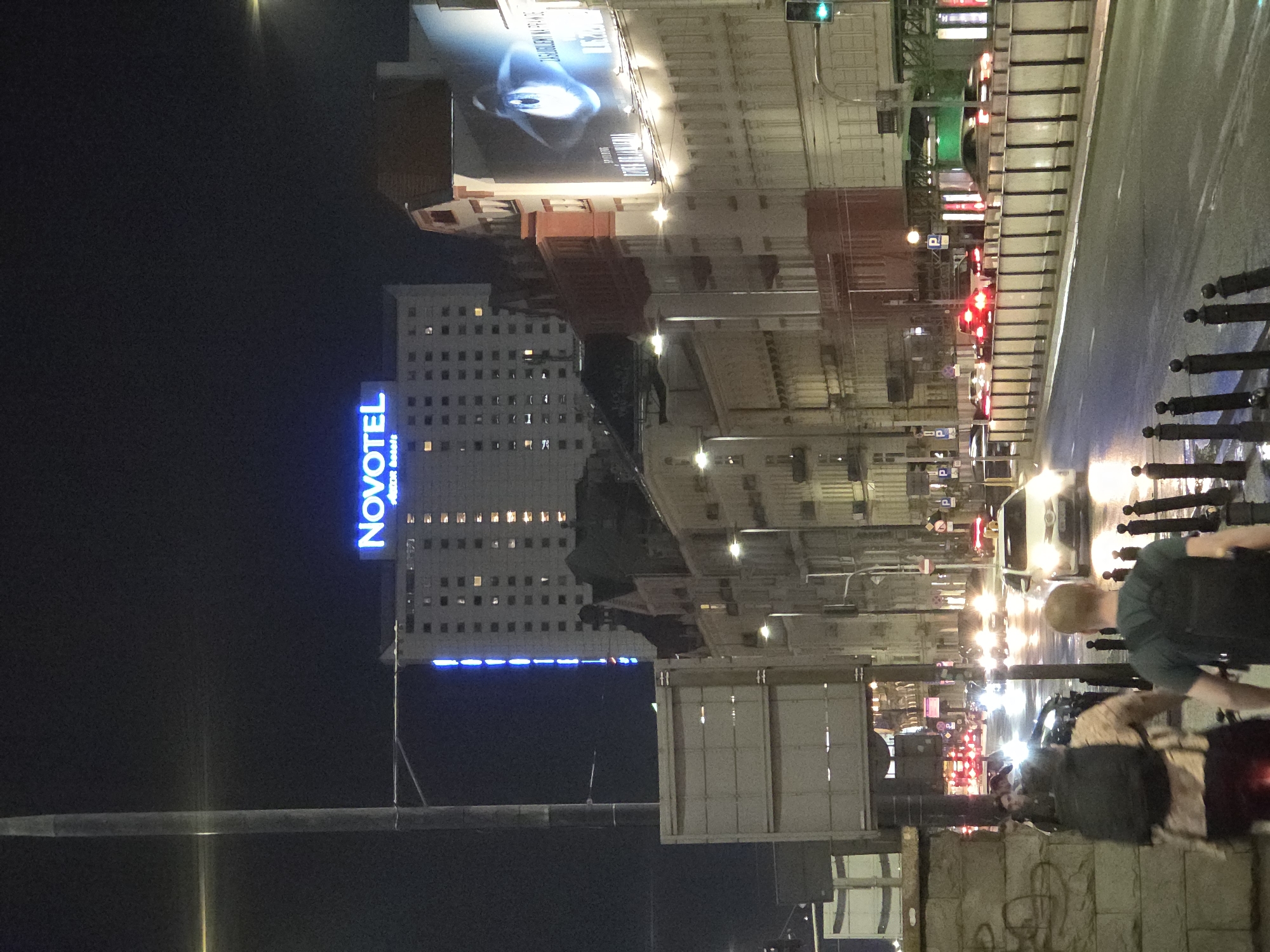 The image shows a night scene in Warsaw, Poland. A Novotel hotel is visible on the left, with its name illuminated in blue. Adjacent to the hotel is a tall building, also lit up with lights. A large billboard is seen above these buildings. On the right, a street with pedestrian traffic, including individuals with backpacks, is visible. Cars with their headlights on are also present, indicating vehicular traffic. Traffic lights are visible, with both green and red lights showing. The scene is lit up by streetlights and vehicle headlights, creating a mix of shadows and bright spots.