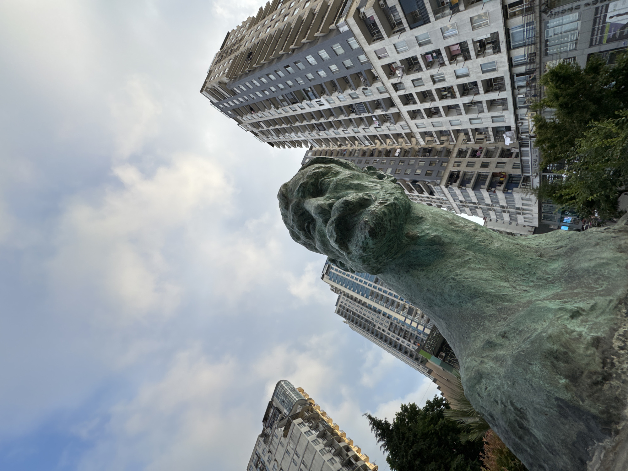 The image, taken from a low angle looking up, captures the statue of a man's head in the foreground, casting a shadow over a portion of the scene. Behind the statue, several tall, modern apartment buildings dominate the skyline. The buildings are characterized by their repetitive rectangular windows and balconies, with some displaying laundry hung out to dry. A portion of a dark green tree is visible on the left side, and some smaller trees and bushes are in the lower right corner. The sky above is partly cloudy with a bright, overcast light, suggesting it might be daytime but not direct sunlight. The overall impression is an urban landscape featuring a prominent sculpture set against the backdrop of contemporary high-rise residential architecture in Batumi, Georgia. The specific location appears to be near the intersection of Gorgasali Street and Memed Abashidze Avenue, as suggested by the presence of the "LEMA" sign on one of the buildings, a known shopping center in the city.