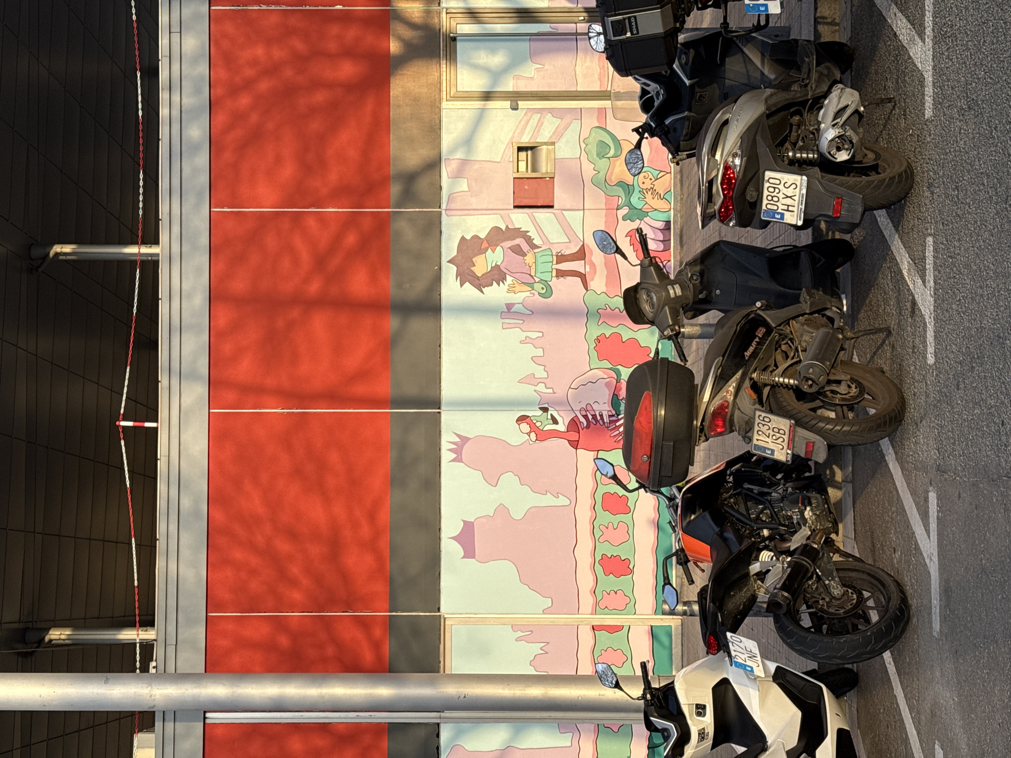 The image depicts a row of parked motorcycles in front of a building with a painted mural. The mural features cartoonish figures and landscape elements in pastel colors. The building facade is divided into sections, some painted red. The motorcycles are parked on a paved surface with painted white parking lines. License plates are visible on the motorcycles. The license plate "0890 HXS" indicates the vehicle is registered in Spain. The license plate "1236 JSB" also indicates the vehicle is registered in Spain. The image is rotated 90 degrees counterclockwise, making the scene appear vertically oriented. I cannot confirm the location is Barcelona, Spain, with certainty without further information.