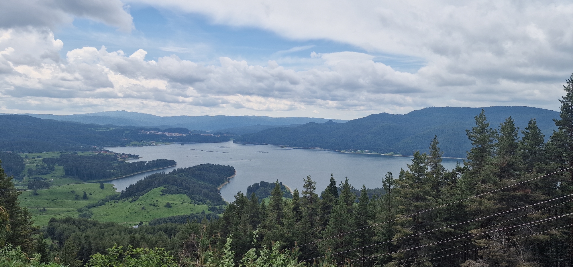 The panoramic image presents a vast and serene mountainous landscape, likely depicting Lake Dospat in Bulgaria. The scene is captured during daytime under a sky dominated by a mix of white and grey cumulus clouds, with some patches of blue visible.
In the foreground, dense evergreen trees, resembling pines or firs, frame the bottom and right side of the view, with some power lines subtly visible through the foliage on the lower right.
The midground is characterized by the expansive, dark blue waters of the lake, which winds through the terrain. Numerous forested peninsulas and islands jut into the water, and on the left, sections of lighter green grassland and more forested hills are visible. Further along the left shoreline, a small cluster of buildings, possibly a village or settlement, can be discerned. Within the lake, several rectangular structures, likely associated with aquaculture such as fish farms, are present.
The background consists of successive layers of undulating mountains, heavily covered in dark green forests, which gradually recede and lighten in color due to atmospheric perspective, extending towards the distant horizon.
The image portrays a tranquil natural environment, devoid of specific human activity beyond the distant settlement and lake structures. No visible text is present. The overall impression is one of natural beauty and an expansive wilderness typical of the Dospat region.