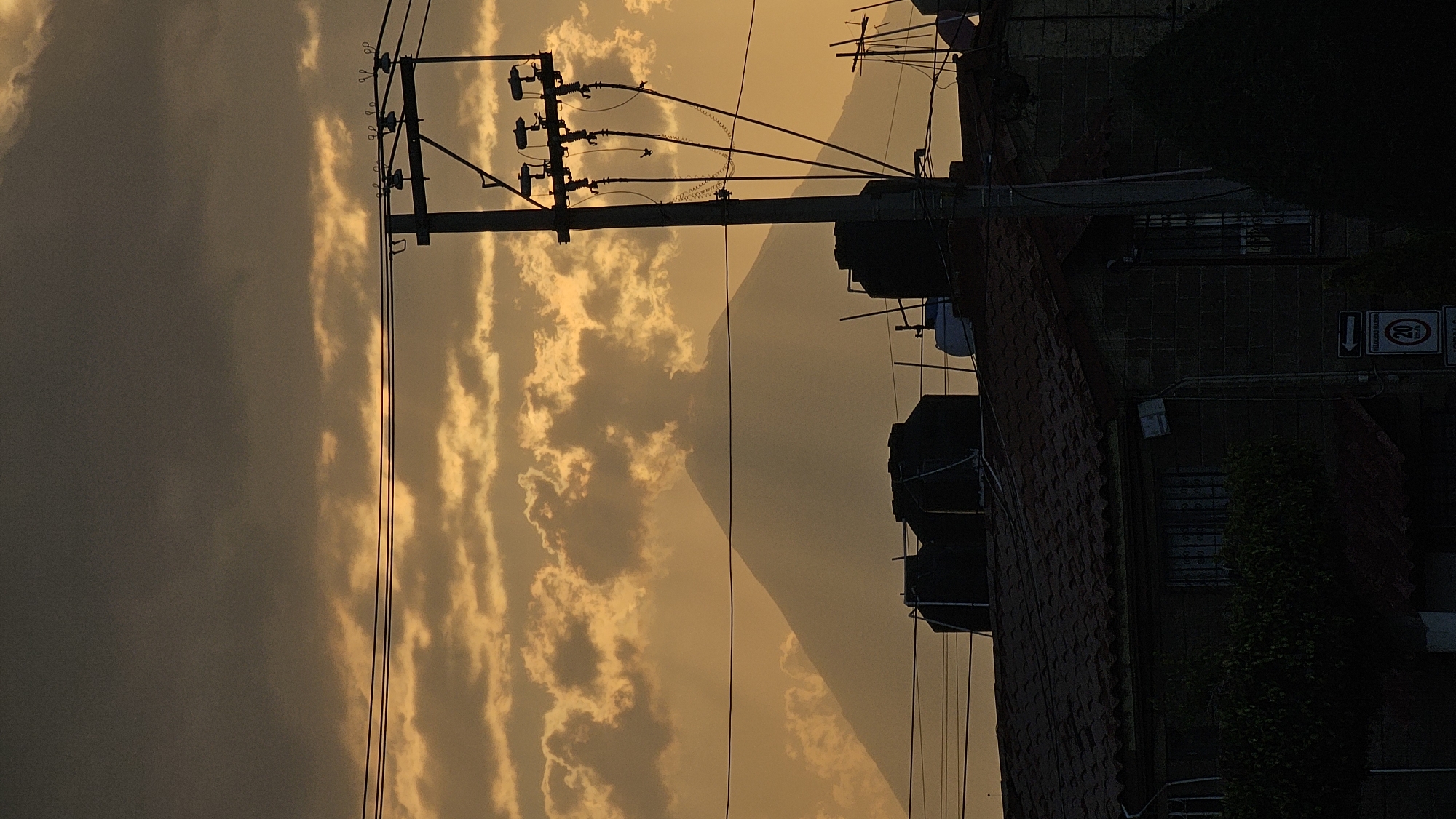 The image depicts an outdoor scene at dusk or dawn in San Bernardino Tlaxcalancingo, Mexico, characterized by a prominent mountain silhouette against a dramatic sky, with urban infrastructure in the foreground.
The upper portion of the sky is dominated by dark, heavy clouds, transitioning to a luminous, golden-orange hue in the lower half where scattered, illuminated clouds are present. A large, conical mountain, likely Popocatépetl, is silhouetted against this bright sky, its peak appearing rounded and with a visible plume of atmospheric haze or ash extending from its upper right flank.
In the midground, a tall, dark utility pole is positioned on the right side, featuring multiple cross-arms equipped with ceramic insulators. A dense network of black power lines extends from the pole and spans horizontally across the frame. Beneath these lines, the dark-tiled rooftops of several residential structures are visible. Multiple dark, cylindrical water storage tanks (tinacos) are mounted on these rooftops, with at least two large ones on the left roof and a lighter-colored, smaller tank with another dark one on the right roof.
The foreground displays the upper facade of a dark-colored building constructed from what appears to be concrete blocks. A window with visible security bars is present on this facade, partially obscured by dark green foliage. In the lower right corner, a white traffic sign with a left-pointing arrow and a red circular emblem indicating a "20 km/h" speed limit is partially visible. No individuals are present within the scene.