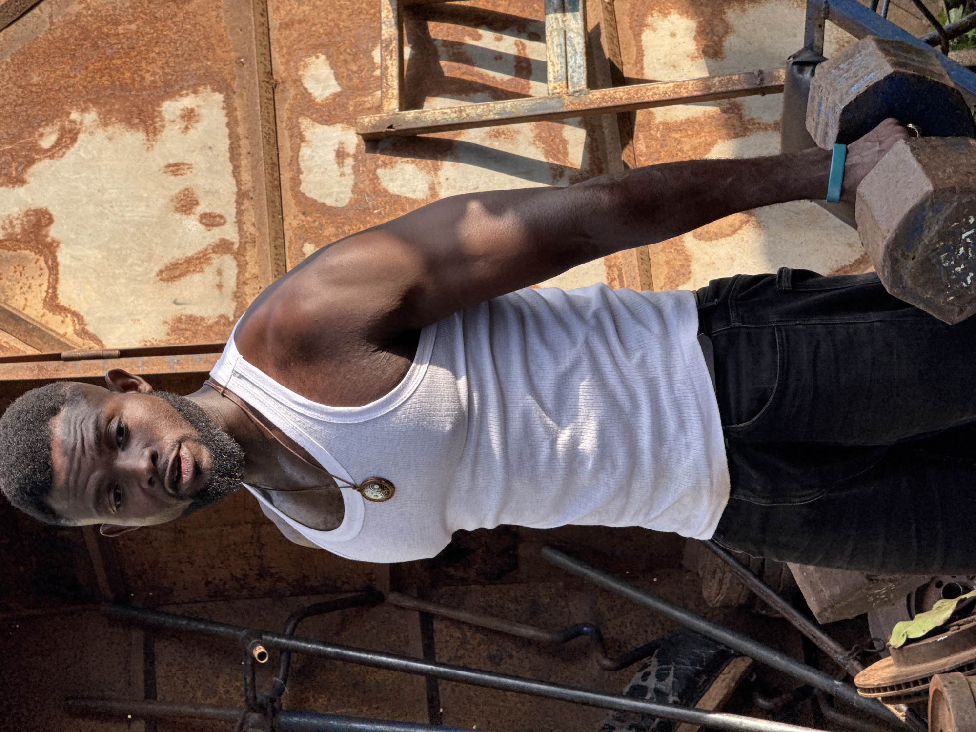 In Akwanga, Nigeria, a Black man stands outdoors, wearing a white tank top and black jeans. He has a beard and short hair, and is wearing a pendant necklace and a turquoise wristband. He is holding a large dumbbell in his right hand, suggesting he is exercising. The background shows a rusty metal structure, possibly a corrugated iron wall, with various metal objects and a ladder leaning against it. The lighting suggests it is daytime, with sunlight casting shadows. The overall scene conveys a sense of gritty urban environment and physical activity.