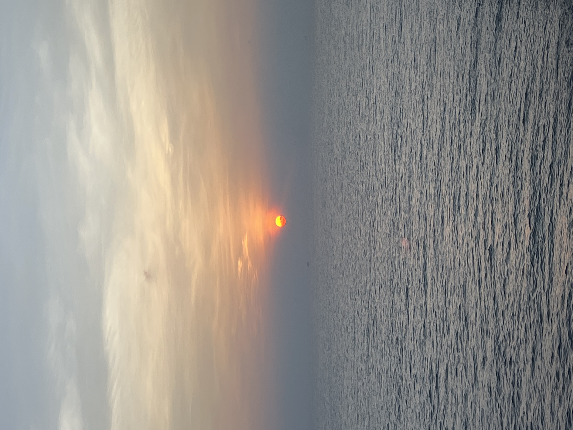 A wide shot captures a serene ocean scene at sunset. The sun, a vibrant orb of orange and red, is partially obscured by wispy clouds, casting a warm glow across the sky. The sky transitions from a pale blue at the top to soft hues of yellow and orange near the horizon, with scattered clouds adding texture. The ocean below is a deep blue, with gentle ripples creating a textured surface. A faint, dark silhouette of a distant vessel is visible on the horizon, barely discernible against the atmospheric haze. The overall impression is one of tranquility and natural beauty. The location is unknown, and no people or specific landmarks are identifiable.