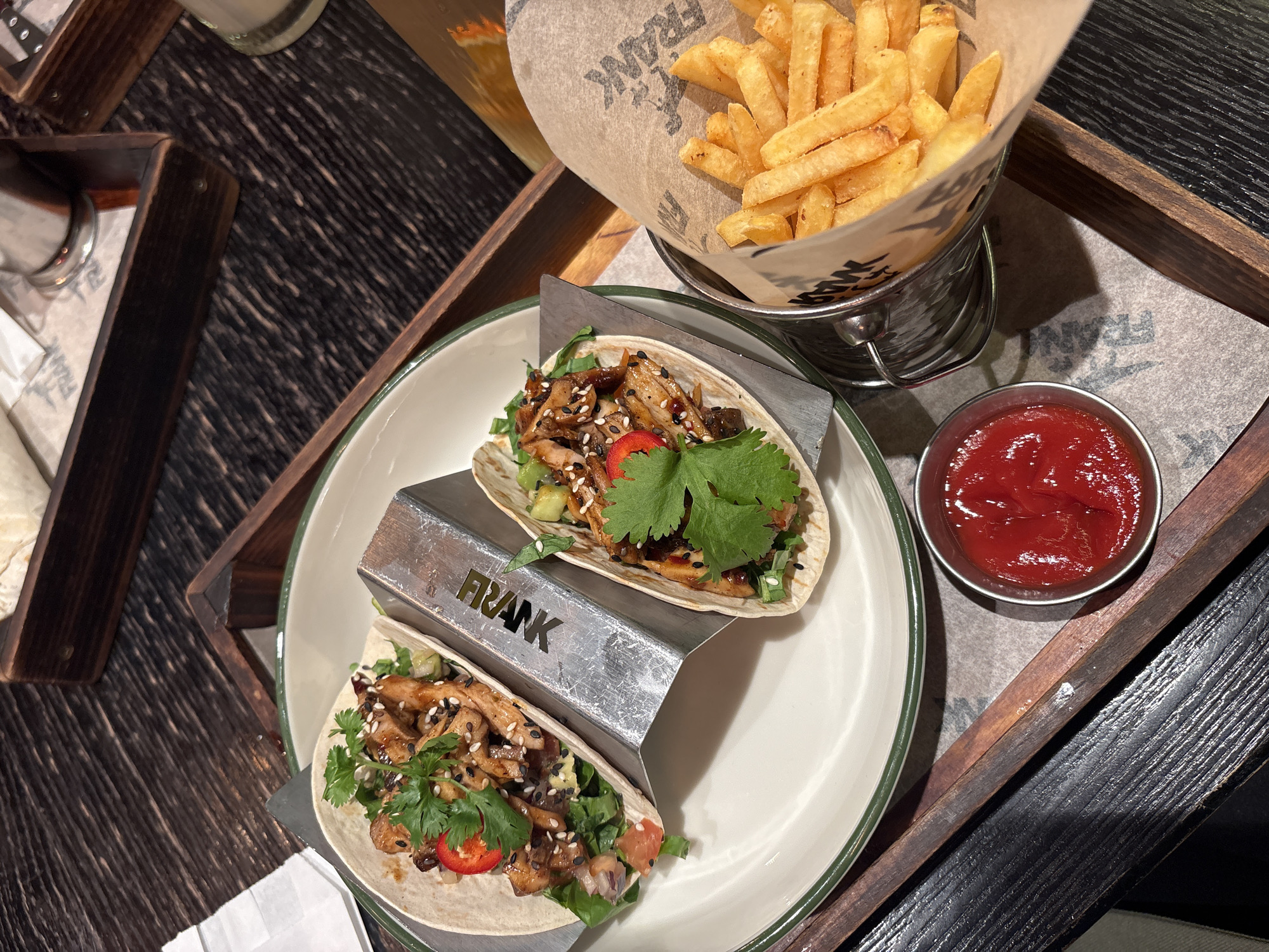 The image shows a close-up of a wooden tray on a dark, possibly wooden, table. On the tray, there is a white plate with two tacos presented on a metal taco holder labeled "FRANK". The tacos are filled with meat, vegetables, and cilantro. Next to the plate, there is a small metal bucket filled with french fries and a small metal bowl containing red sauce. A glass is visible in the upper left of the frame, and a bottle is in the background behind the fries. The overall scene suggests a casual dining experience, likely in a restaurant. The presence of the "FRANK" branding suggests this is the name of the establishment. While the city of Saint Petersburg, Russia, cannot be confirmed without more context, the image depicts food service and presentation common in urban restaurant settings.