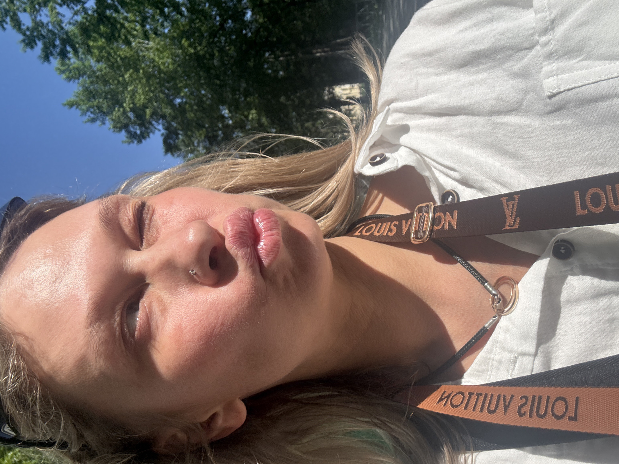 A close-up photograph shows a fair-skinned woman with blonde hair lying on her back outdoors, with a tree and blue sky visible in the background. The woman has a nose piercing and pursed lips. She is wearing a white collared shirt and a Louis Vuitton lanyard or strap around her neck. The image appears to be taken during the day in a sunny location. There is no information to deduce that the photo was taken in the city of Perm, Russia.