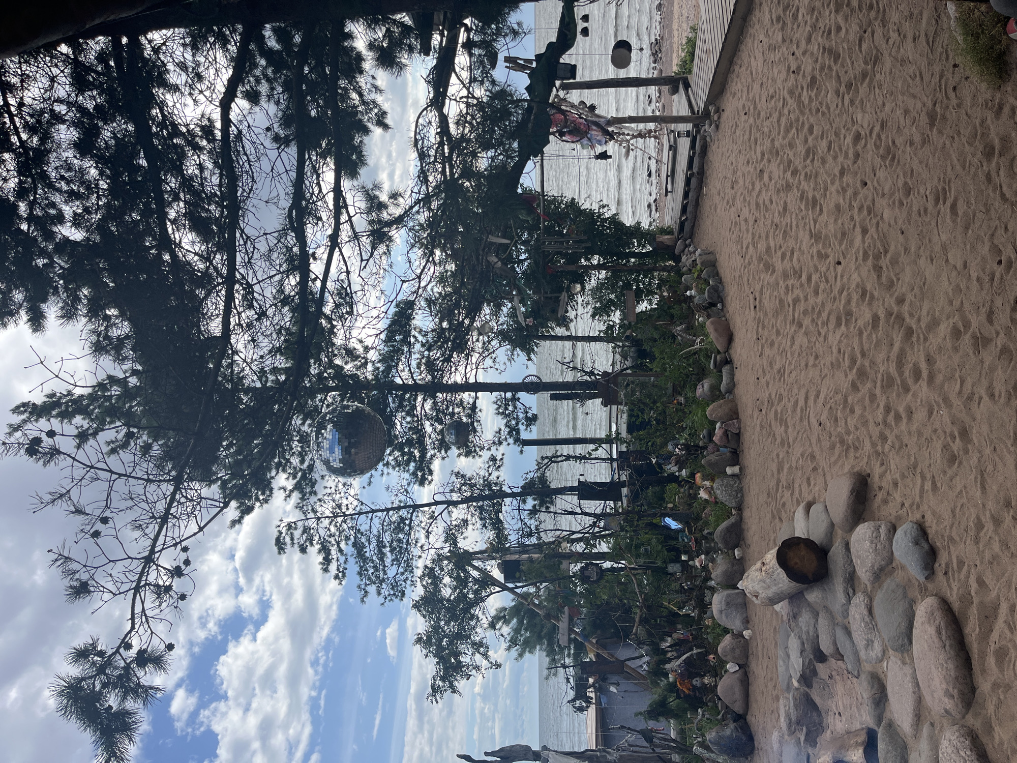The image depicts a sandy area with a fire pit made of stones and a log, located near a body of water. Pine trees are visible in the foreground and midground, with a disco ball hanging from one of the branches. The trees are decorated with various ornaments and objects, creating a cluttered, artistic display. In the background, the sea stretches to the horizon under a cloudy sky. A wooden walkway leads to a platform near the water, with more decorations hanging from structures. The scene appears to be outdoors, possibly a beach or a coastal area, with a bohemian or eclectic ambiance. There are no people visible in the image.