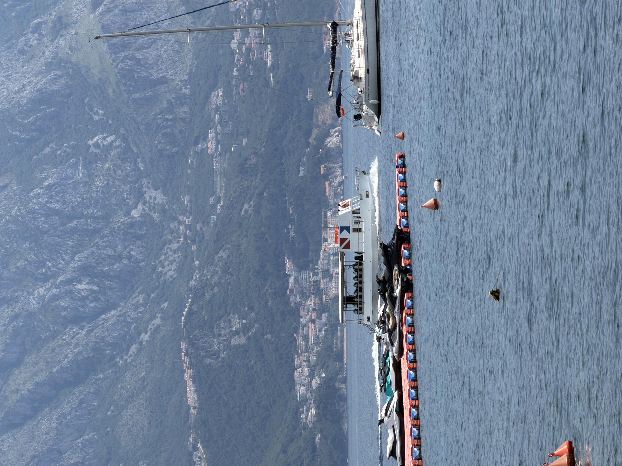 The image shows a waterfront scene in what appears to be the city of Budva, Montenegro. In the background is a steep, mountainous terrain, dotted with clusters of buildings, likely residential areas. In the foreground, a body of water takes up a significant portion of the scene, with visible ripples on the surface. A large vessel, possibly a passenger ferry or a tour boat, is positioned parallel to the shoreline. It's laden with watercraft, primarily jet skis and other water sports equipment, secured along a floating dock made of orange and blue pontoons. Near the boat, there's a sailboat. Scattered throughout the water, there are a few navigation markers in bright orange and a single seagull.