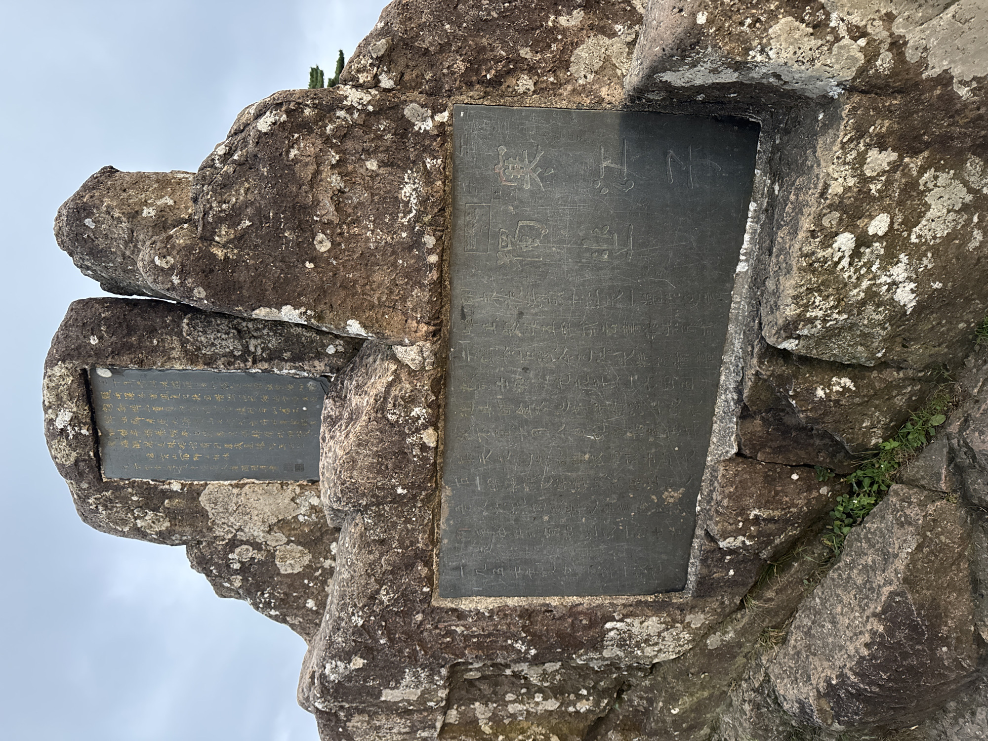 A tablet with Chinese calligraphy is embedded in a large rock formation. The tablet is rectangular and dark, with the characters carved into its surface. The rock formation is rough and textured, with patches of lichen growing on it. The sky in the background is overcast. The image is taken from a low angle, looking up at the tablet and rock. The scene is located in the city of Huangshan District, China.