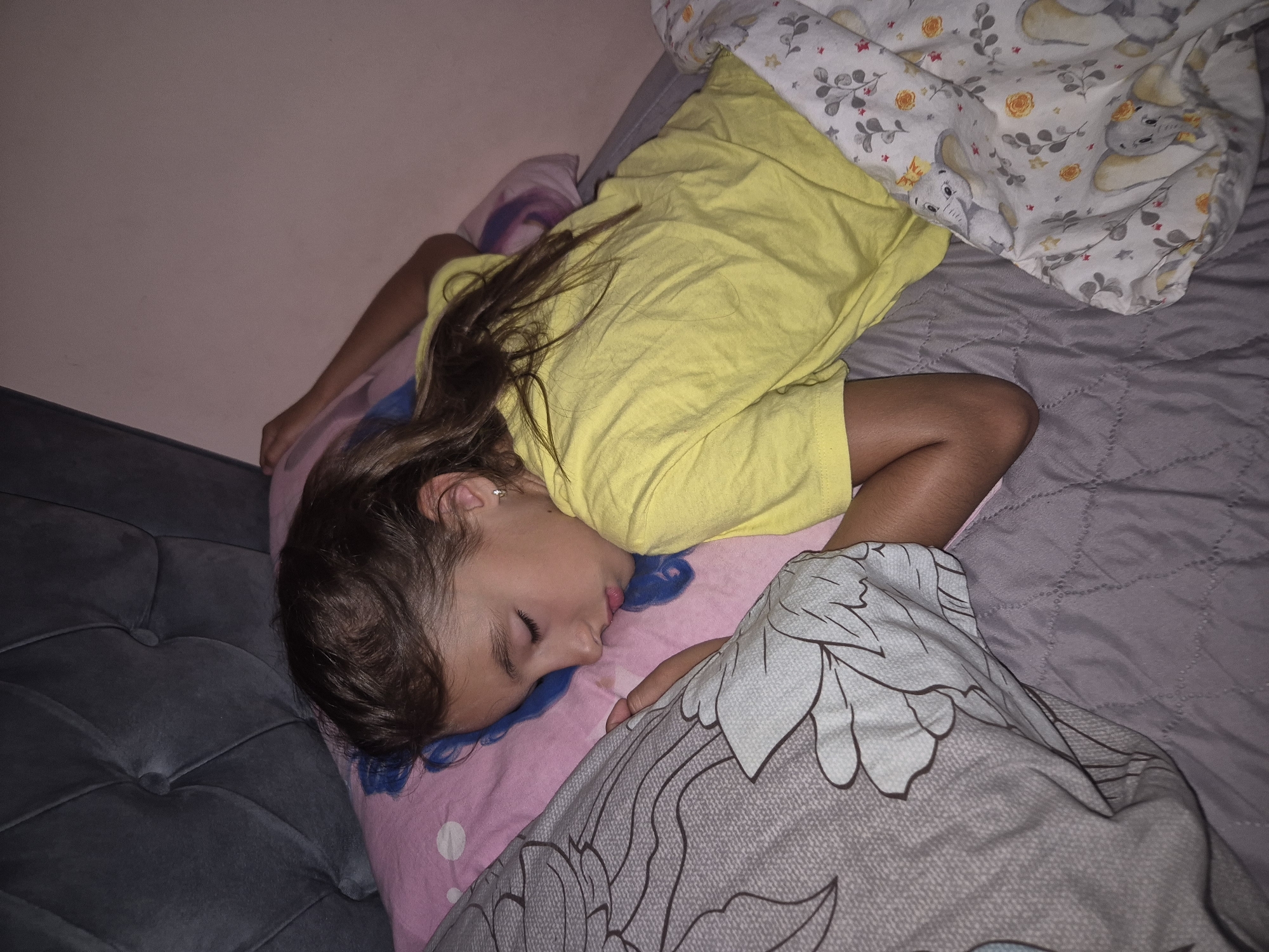 A young girl is sleeping on her side, facing the camera. She is wearing a yellow t-shirt and is covered by a gray blanket with a floral pattern. The girl is lying on a pink pillow with polka dots. Her hair is long and brown, and it is spread across the pillow. Her eyes are closed, and her mouth is slightly open. The background is a pink wall. The scene appears to be indoors, likely a bedroom. The lighting suggests it might be nighttime or a dimly lit room. The overall mood is peaceful and serene, as the girl is deeply asleep. There is no visible text in the image.