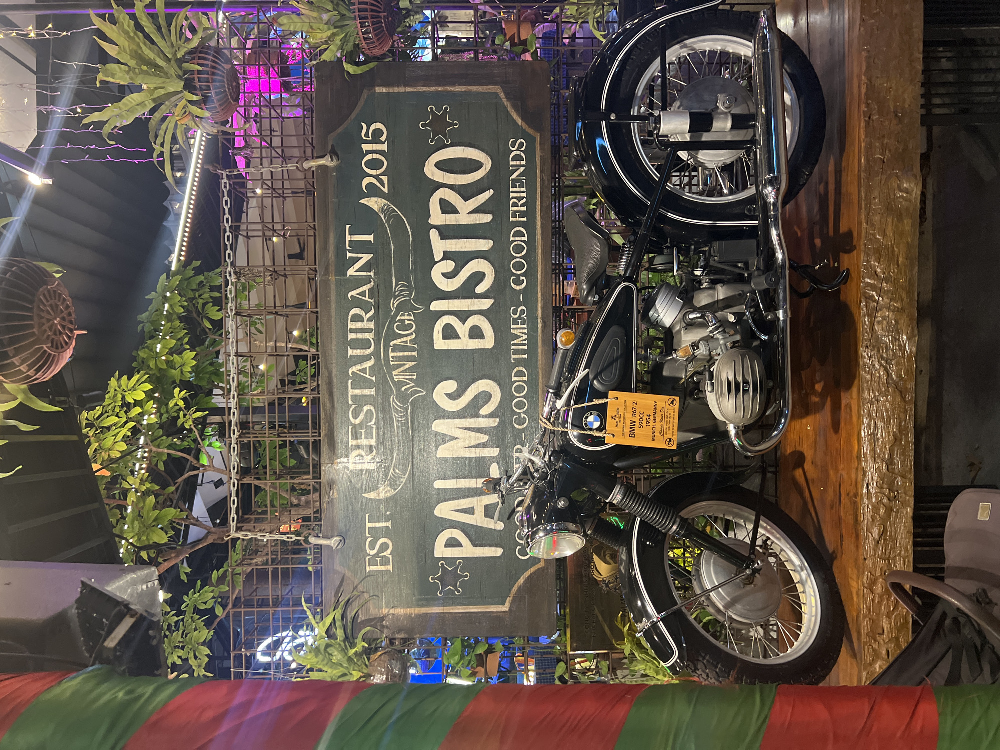 An antique black BMW R67/2 motorcycle is displayed prominently on a wooden surface, likely a bar or restaurant counter, in Nha Trang Ward, Vietnam. The motorcycle, a 1954 model with a 590cc engine, is positioned in front of a dark green wooden sign that reads "EST. RESTAURANT VINTAGE PALMS BISTRO 2015 GOOD TIMES - GOOD FRIENDS". The sign features stylized lettering and decorative elements, including a sheriff's star. A yellow tag is attached to the motorcycle's handlebars, detailing its make and model. The background consists of a dimly lit interior with hanging plants, decorative lighting, and metal fencing, suggesting a restaurant or bar setting. A colorful striped pillar is visible to the left of the motorcycle.