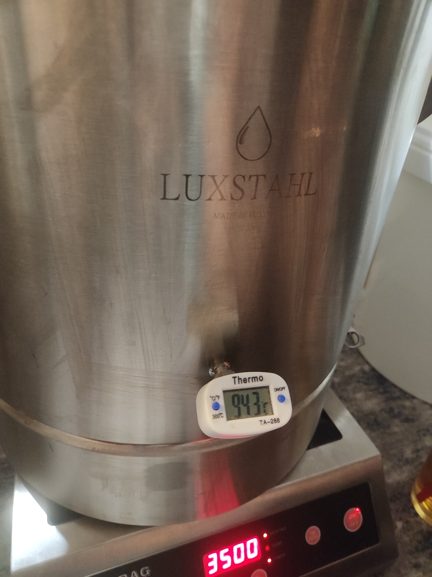 A close-up shot focuses on a stainless steel container, likely a brewing or cooking vessel, displaying the brand "LUXSTAHL" and a droplet logo on its surface. A digital thermometer attached to the container reads a temperature of 94.3 degrees. The vessel sits atop a digital scale, which displays the weight "3500".