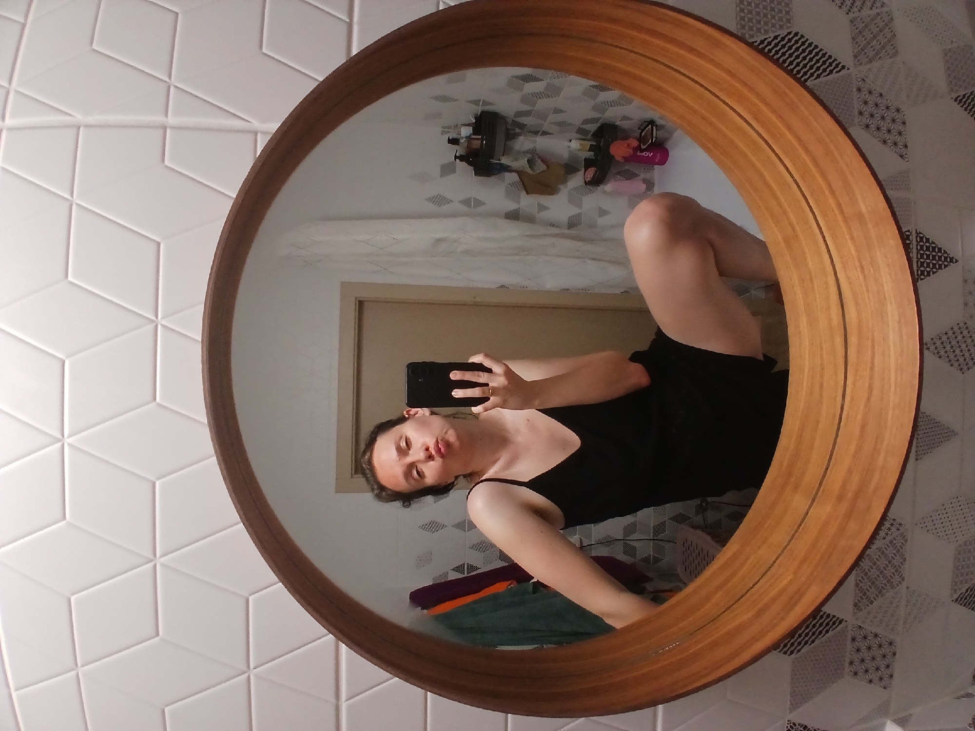 In Yekaterinburg, Russia, a woman is taking a selfie in a round wooden-framed mirror in a bathroom. She is wearing a black camisole and shorts, with her right leg bent and her left leg extended. Her lips are pursed, and she is holding a black smartphone in her right hand. The bathroom features white geometric tiles on the wall and a patterned tile backsplash. In the reflection of the mirror, various bathroom items are visible, including towels, bottles, and shelves.