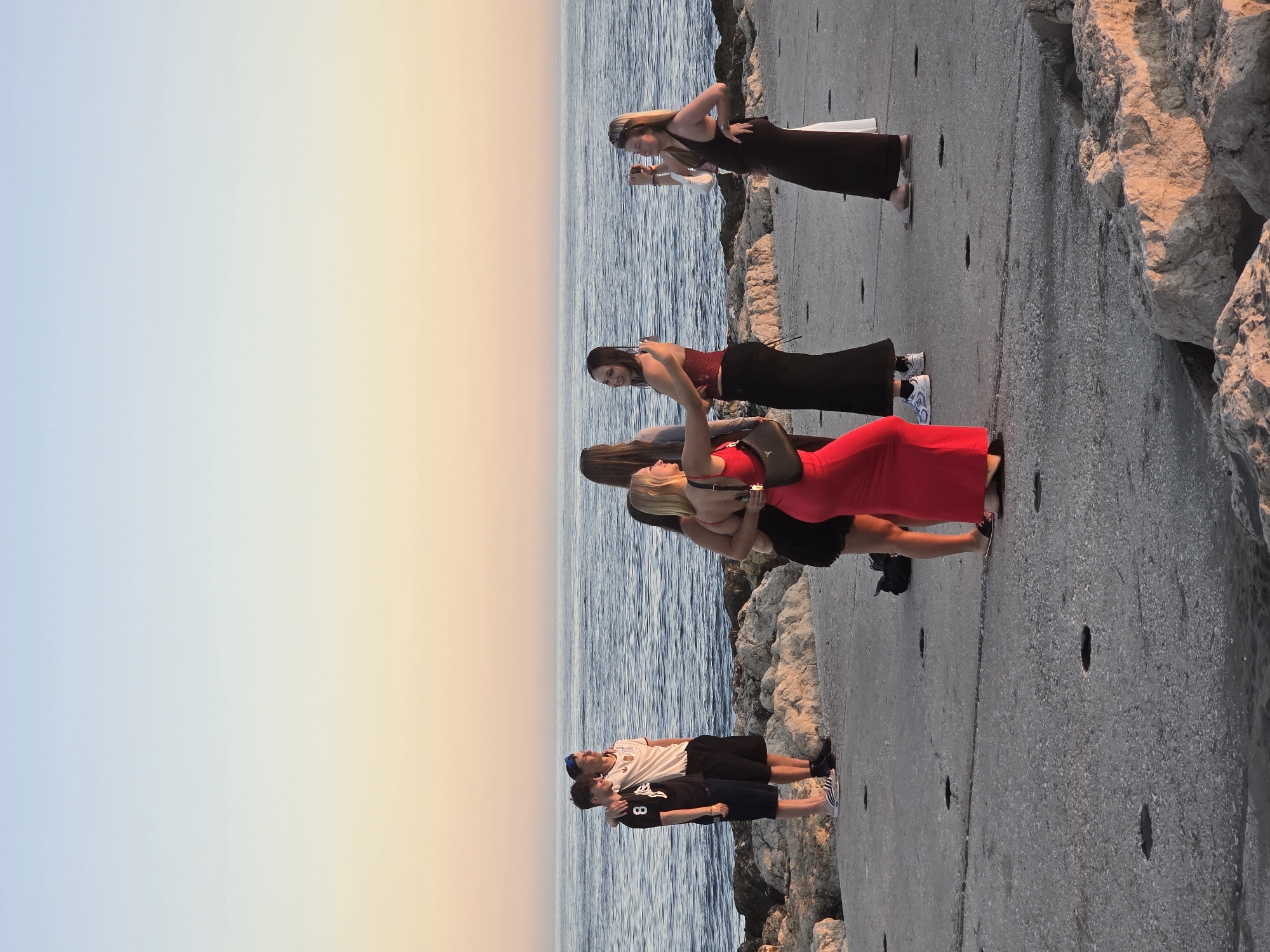 The image depicts a group of young adults gathered on a paved promenade along the coast. The scene appears to be set at sunset, with the sky displaying a gradient of pale blue to warm orange and pink hues. The sea is calm and stretches to the horizon. In the foreground, there are large rocks, typical of the coastline in Málaga, Spain.
On the promenade, two young men are standing together, with one man's arm around the other's shoulder. They are both casually dressed. Further along, a group of young women are interacting. One woman in a vibrant red dress is posing for a photo, while another woman in a black dress is taking her picture with a smartphone. Two other women, one in a dark top and black skirt, and another in a white top and dark skirt, are also present and appear to be engaged in conversation. The overall atmosphere suggests a social gathering, possibly before or after an event, enjoying the seaside scenery. The lighting indicates it is late afternoon or early evening.