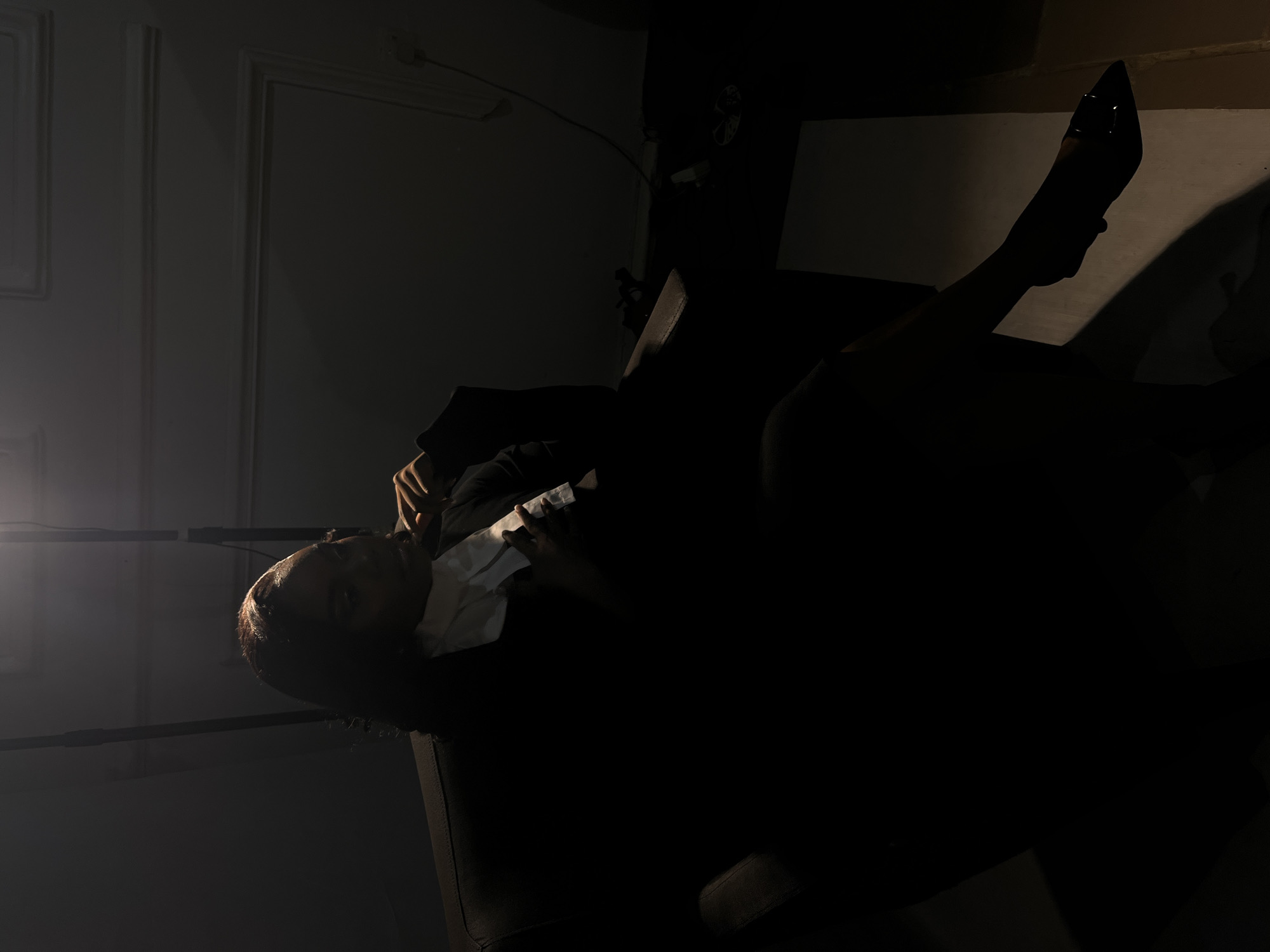 A dark, high-contrast image shows a woman in a dimly lit room, possibly an office or studio. She is seated in a dark chair, leaning forward with one hand resting on her chin and the other on her chest. She appears to be wearing a white collared shirt or blouse underneath a dark jacket or suit. The lighting is dramatic, with a spotlight casting shadows that obscure much of the scene. The background shows a wall with decorative molding, and to the right, some electrical outlets and cords are visible. The overall impression is one of contemplation or introspection.