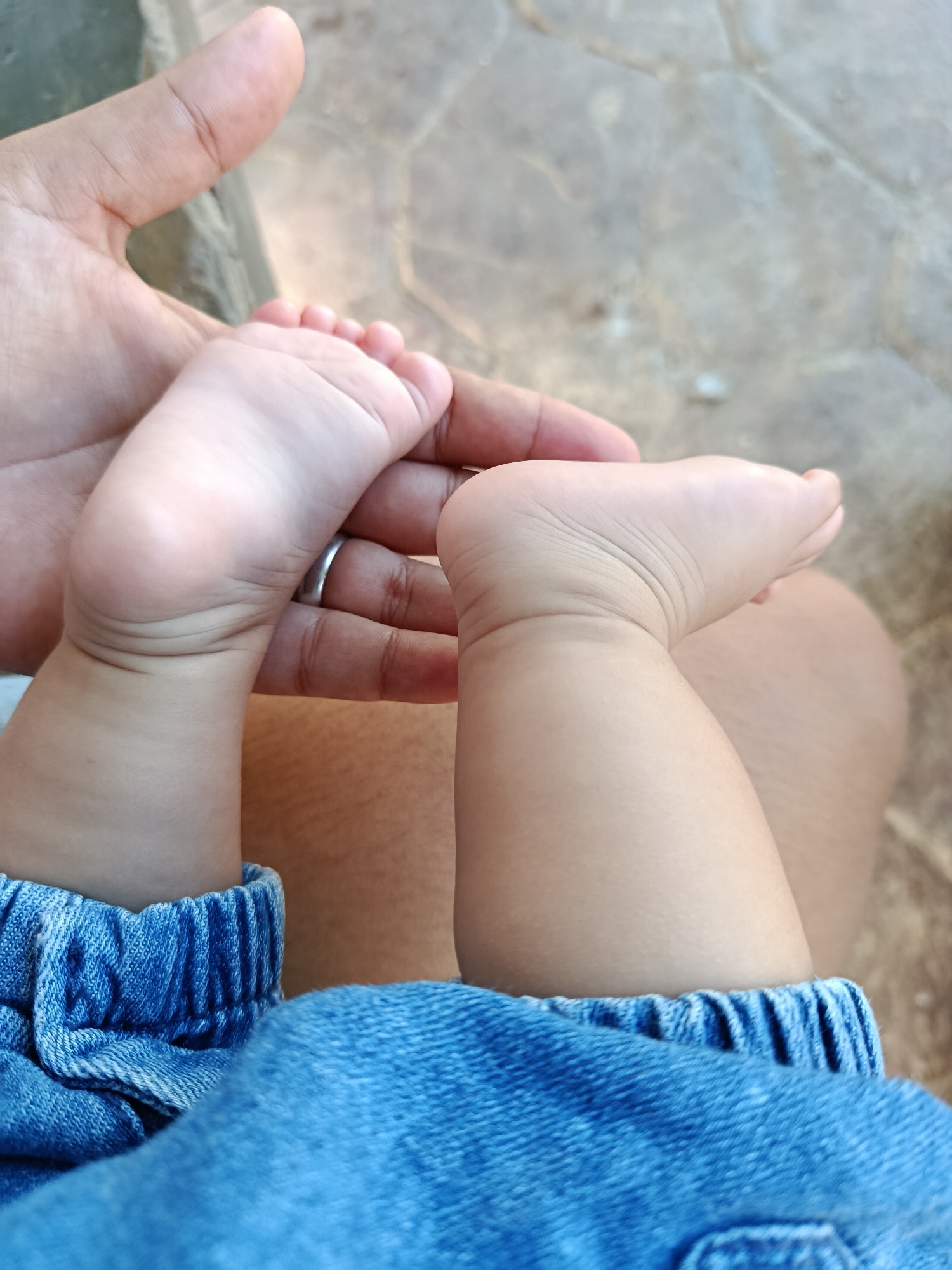 The image shows a close-up view of a baby's feet and an adult's hand. The baby's feet are small and delicate, with visible toes and soft skin. A silver ring is worn on the adult's finger. The baby is wearing blue denim shorts with an elastic waistband. The background is blurred, showing what appears to be a stone or tiled surface, possibly an outdoor patio. There are no clear indicators within the image to suggest a specific location in Juan Griego, Venezuela. The lighting suggests an outdoor setting during the day.