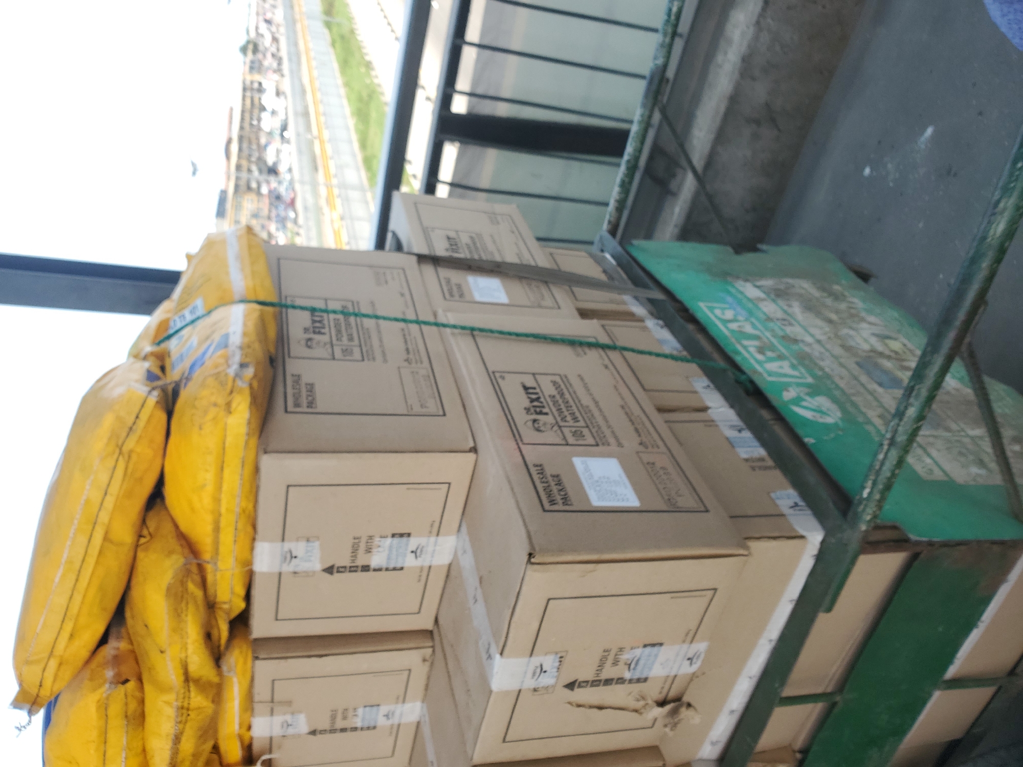 A green metal trolley is heavily loaded with packages, situated in an outdoor or semi-outdoor area, likely a loading dock or transport hub in Lagos, Nigeria. The trolley carries a substantial stack of brown cardboard boxes on its right side, secured with white tape and a green rope. Visible text on the boxes includes "HANDLE WITH CARE" (indicated by upward arrow symbols), "WHOLESALE PACKAGE," "FIXIT," "POWDER," and "106 WATERPROOF." On the left side of the trolley, several large, bright yellow sacks or bags are piled, also secured by a green strap.
In the background, a brightly lit urban scene unfolds under clear skies, suggesting daytime. A busy road or street is visible in the distance, populated with numerous vehicles, including cars and motorcycles, characteristic of a bustling city. An elevated structure or overhead walkway runs parallel to the road. A partially visible green sign or tarp on the right side of the trolley displays the word "ATLAS." The arrangement suggests a logistical operation, such as the loading or preparing of goods for transport or delivery.