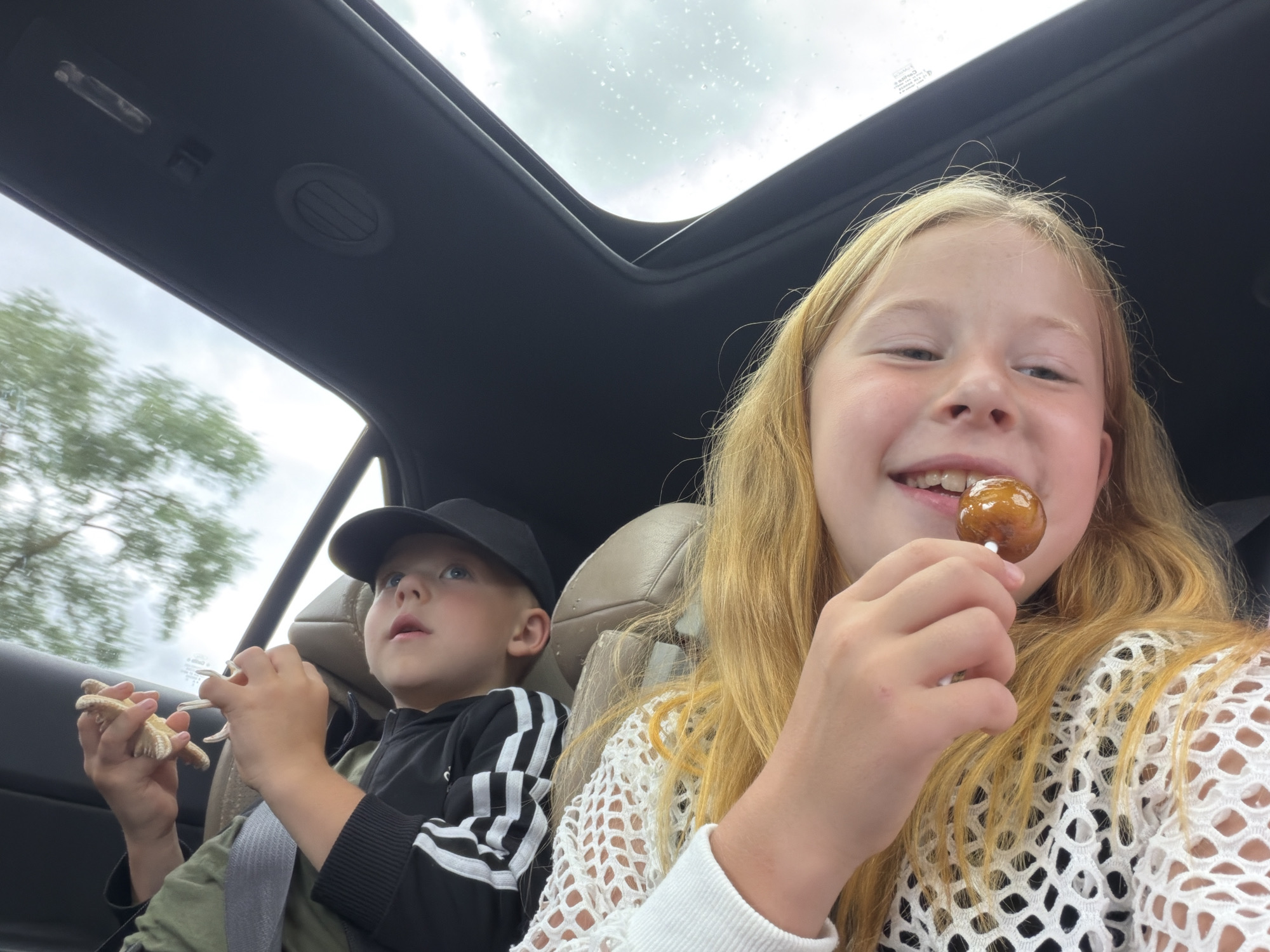 Two children are seated in the backseat of a car, with a sunroof open to a cloudy sky. The child on the right, a girl with long blonde hair, is smiling and holding a lollipop near her mouth. She is wearing a white crocheted top over a black garment. The child on the left, a boy wearing a black baseball cap and a black jacket with white stripes, is holding two toy starfish. The interior of the car appears to be leather. The background shows blurred trees, indicating the car is in motion, likely on a road.
