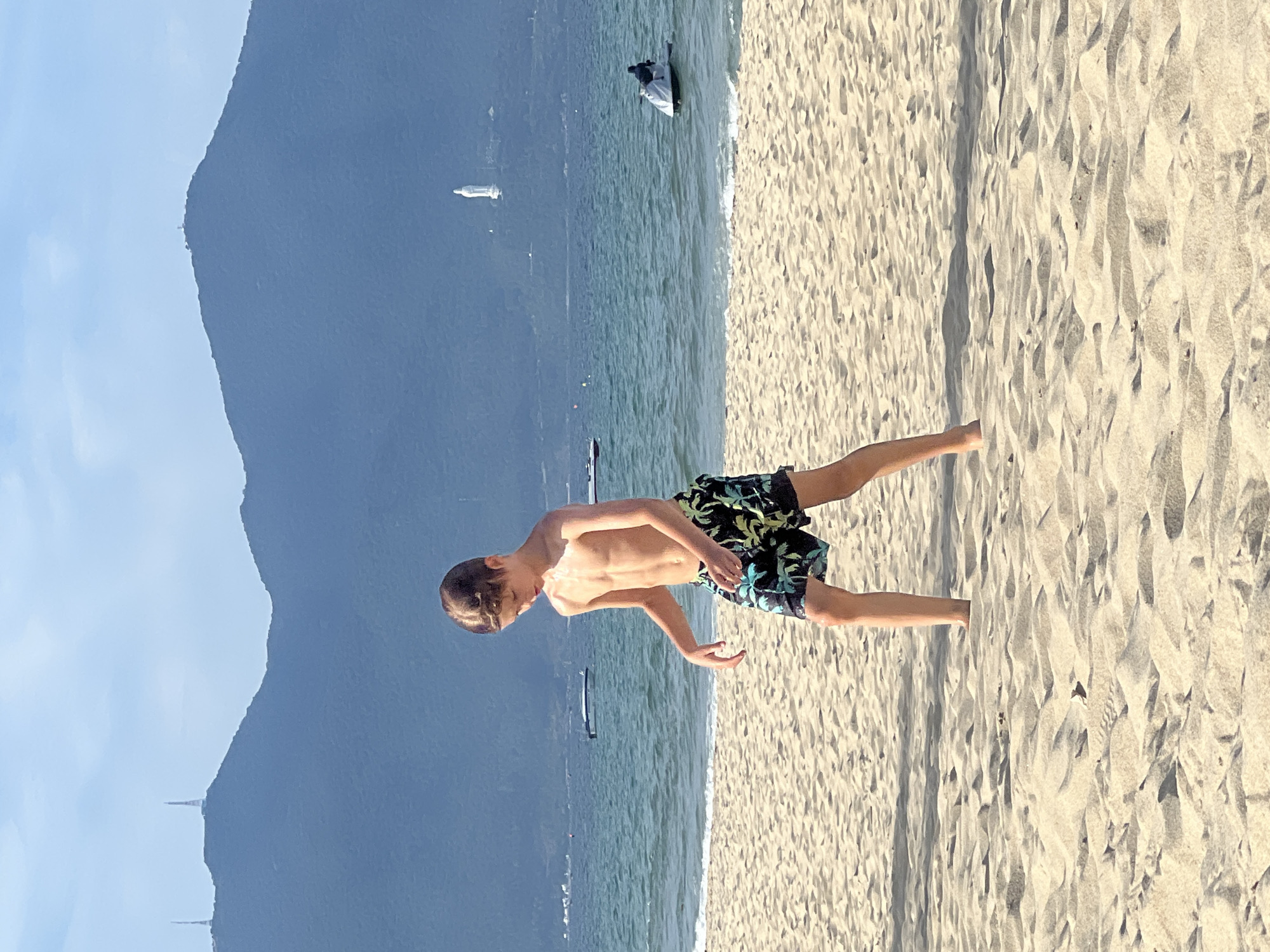 A young boy with fair skin and light brown hair, wearing black and green palm-tree patterned swim trunks, stands on a sandy beach. He is shirtless and appears to be walking or looking down. His left leg is slightly in front of his right. The sand is light beige and textured with ripples. In the background, a calm, turquoise ocean stretches towards a hazy, blue mountain range under a light blue sky with scattered clouds. Two antennas are visible on the leftmost mountain peak. Further to the right, a white statue of a person is perched on the side of a mountain. Several boats are scattered on the water, and a person is riding a white jet ski in the middle ground. The scene is likely located in Da Nang, Vietnam, given the prominent coastal landscape and the statue, which resembles the Lady Buddha statue on Son Tra Peninsula.