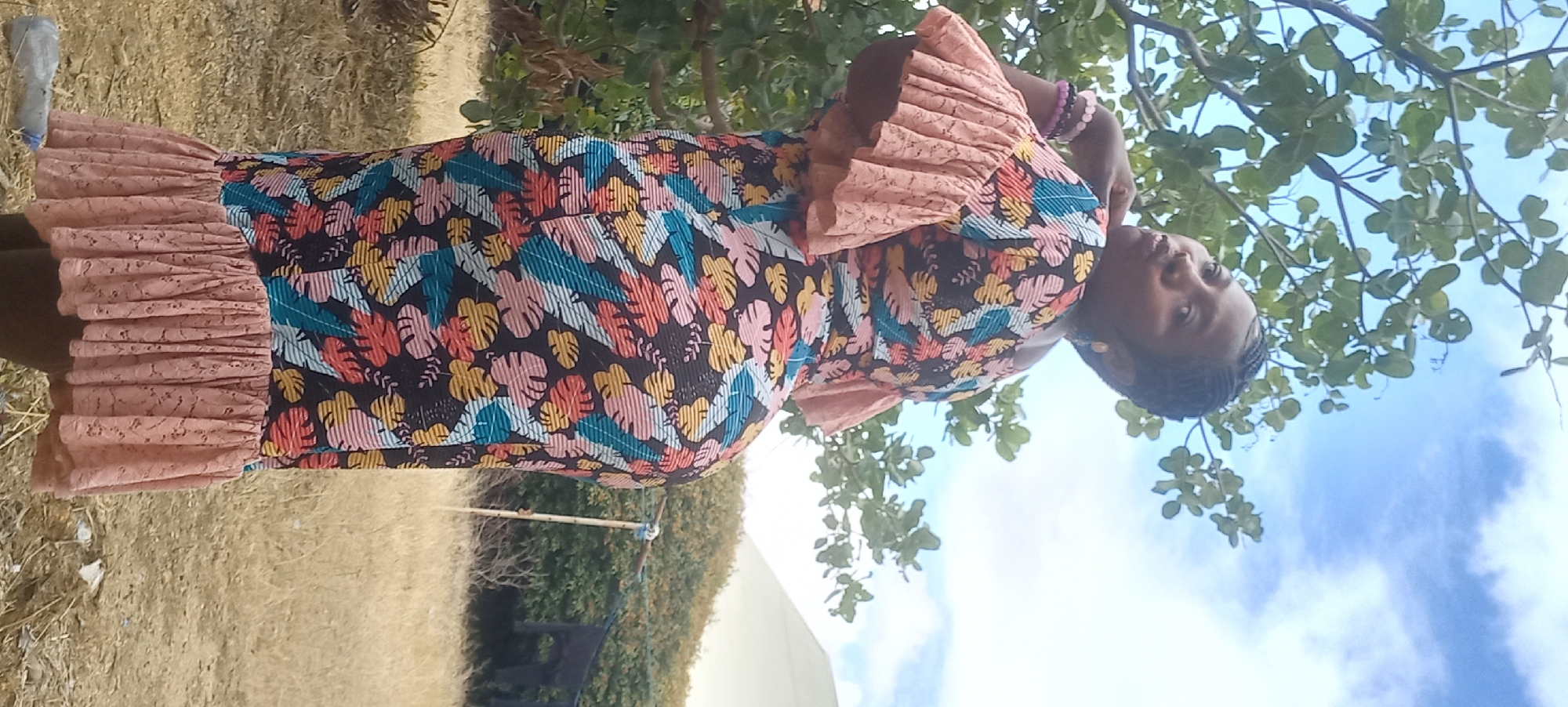 A woman stands outdoors, her body angled away from the camera, with her head turned to look back over her shoulder. She is wearing a vibrantly patterned dress with a floral design. The dress has ruffled short sleeves and a ruffled hem. She is adorned with bracelets on her left arm. The background consists of a tree with green leaves, a cloudy blue sky, dry grass, and some shrubs. In the distance, a pale dome-shaped structure is visible, along with a clothesline and some dark fabric hanging on it. The environment appears to be a somewhat dry, outdoor space, possibly a yard or open field. The overall impression is a casual, outdoor portrait. There is no identifiable information about the location being Jos, Nigeria.