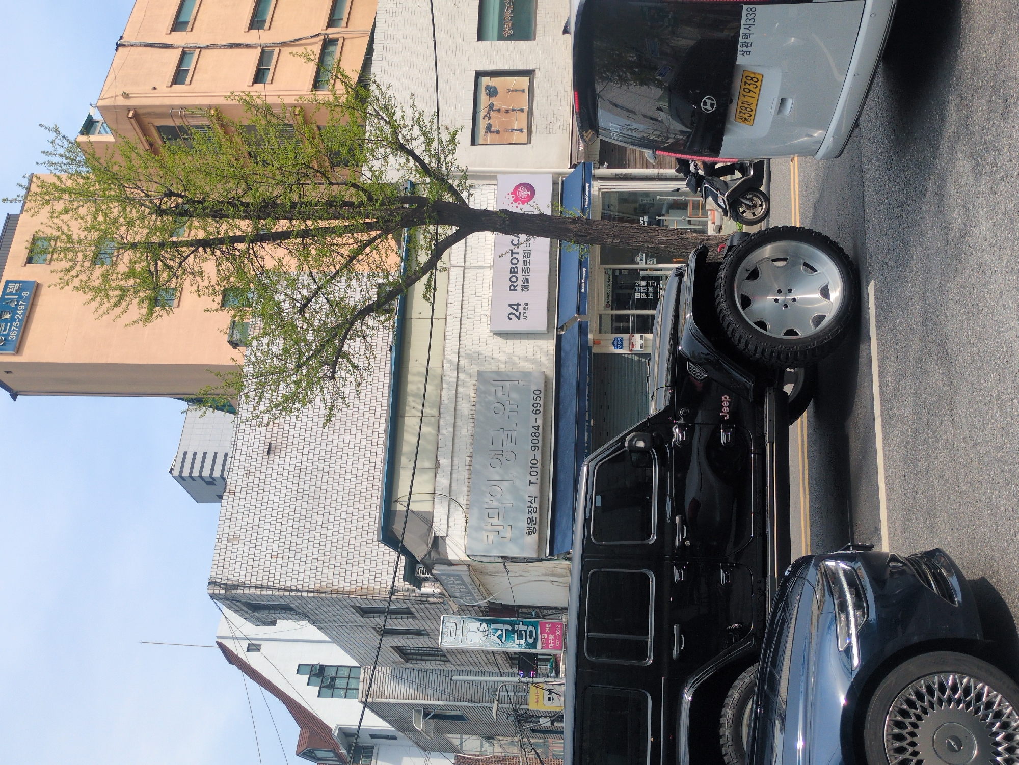 The image depicts a street scene in Seoul, South Korea. The view is from ground level, looking across the road towards a building with signage in Korean. A tree with sparse green leaves partially obscures the building. Signage visible includes "24 ROBOT CA 해솔(종로점) be" and "칸막이 앵글 유리." Additional signs offer services like "아구찜 대구탕 전문." A black Jeep with a silver spare tire is parked on the road, partly blocking the view of the building. A dark blue sedan is parked closely in front of the Jeep. A white van and a motorcycle are parked behind the Jeep across the street. The license plate "설38자 1938" can be seen on the white van. The buildings in the background consist of a mix of styles, with one being a salmon-colored high-rise structure.