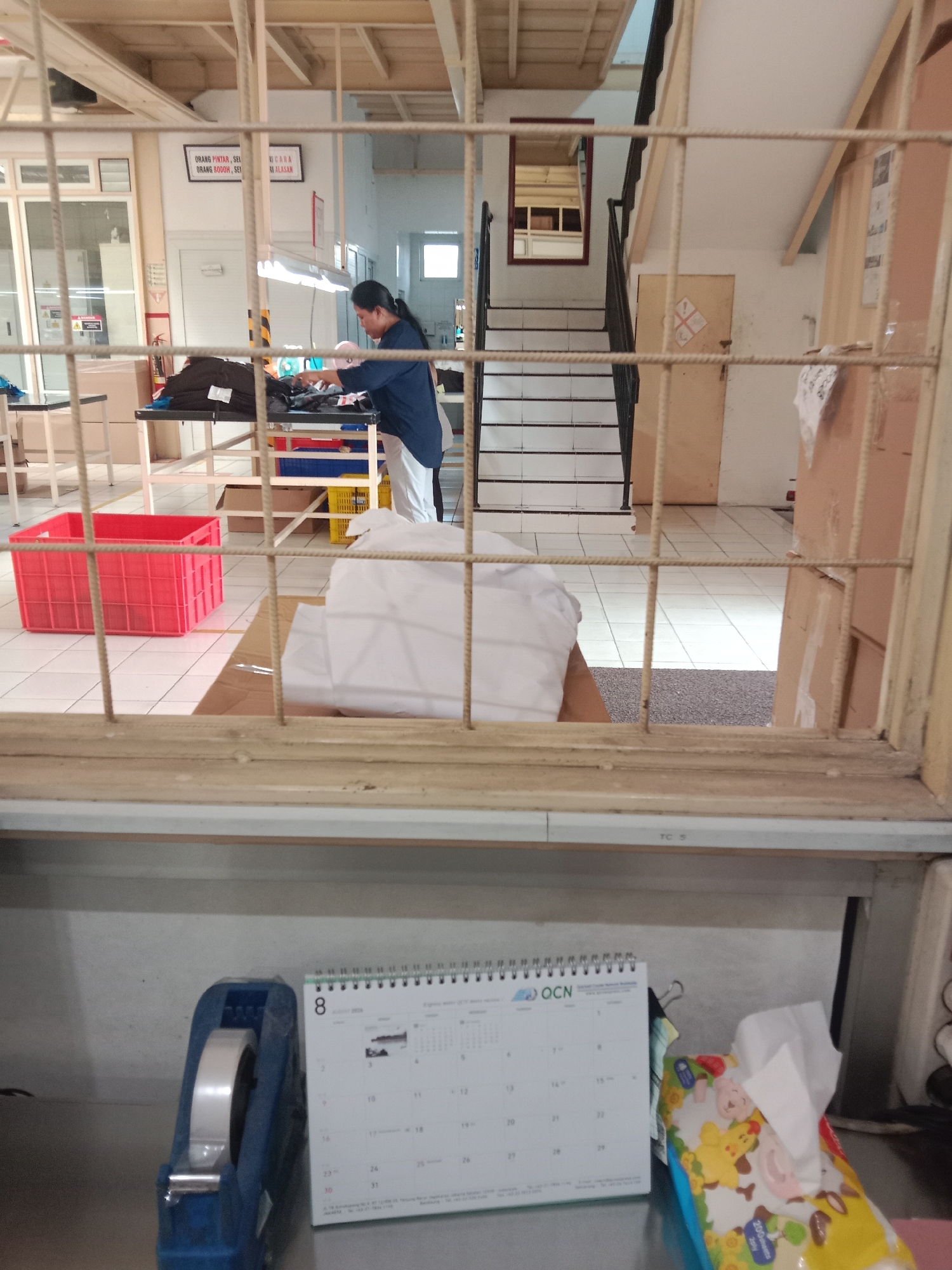A woman in a blue shirt and white pants is folding white cloth on a table in what appears to be a factory or warehouse. The scene is viewed through a metal grate. To the left, a red plastic bin is visible, and in the background, a staircase with a mirror at the top can be seen. On a shelf in the foreground, a tape dispenser and a calendar are present. The environment suggests an industrial setting within a city.