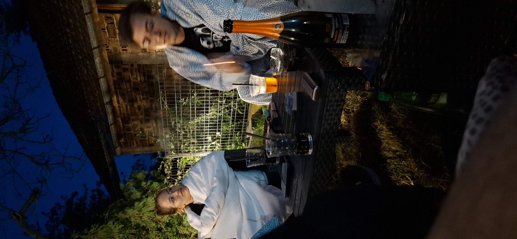 The image depicts an outdoor gathering at dusk or night, likely in the vicinity of Lobosh, Bulgaria, given the context provided. Two individuals are seated at a dark-colored, woven outdoor table. The person on the left, a woman with glasses, is wrapped in a white blanket and looking to her right. The person on the right, a man wearing glasses and a black t-shirt with a graphic print, is also covered by a patterned blanket. He appears to be holding a straw in a drink, and a faint orange glow, possibly from a light source or a cigarette, is visible near his hand.
On the table, there are several items: two glasses with dark liquid and straws, a cup with an orange beverage and a straw, a cigarette lighter, what appears to be a pack of cigarettes, an ashtray, and a bottle of sparkling wine with an orange label. The background shows a building with a brick wall and a dark roof, a metal fence, and lush green foliage, including tree branches silhouetted against the dark blue night sky. The ground in front of the table is covered with grass. The overall atmosphere suggests a relaxed social gathering.