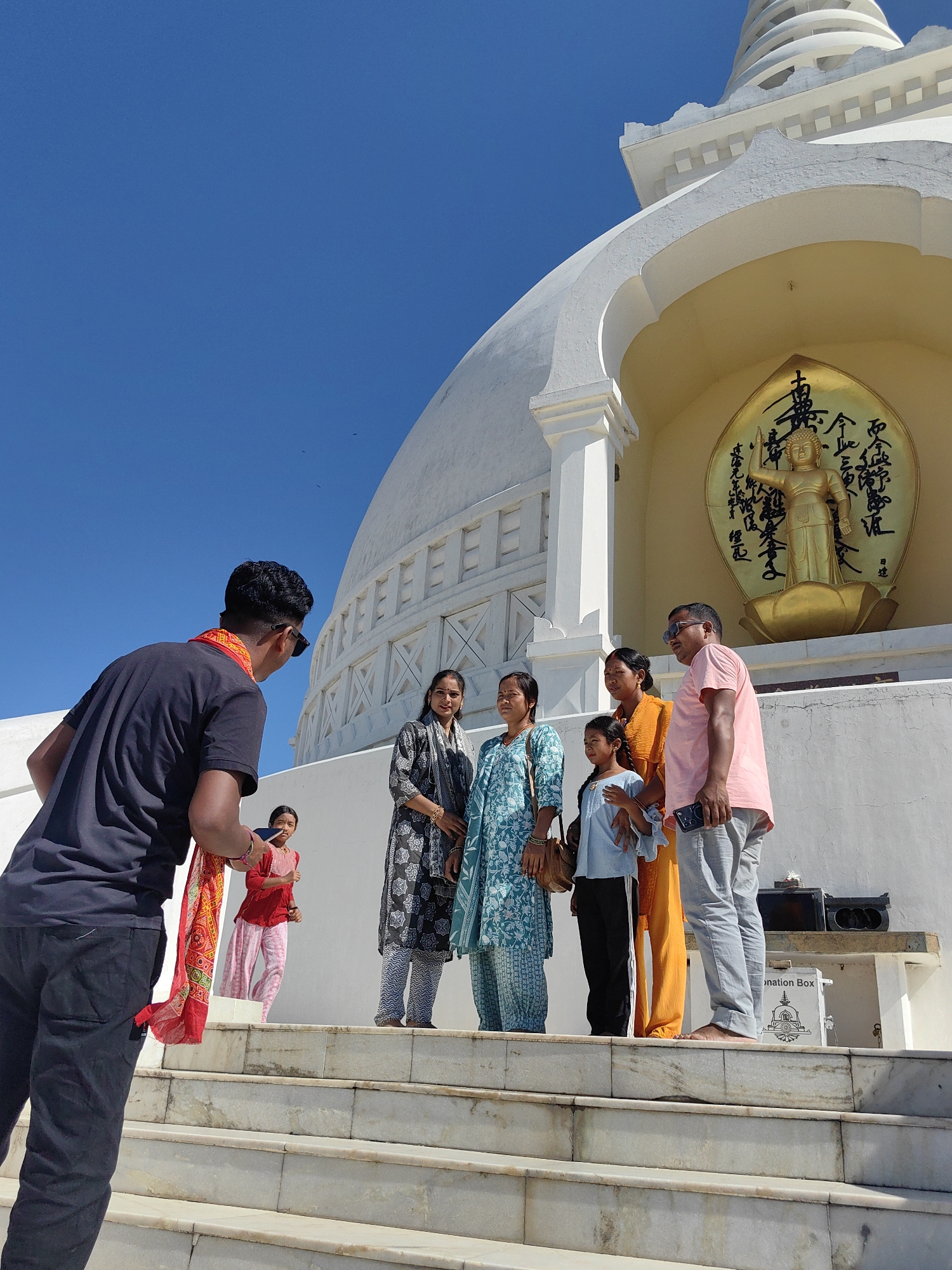 The image depicts a bright, sunny day with a clear blue sky, likely in Parsa, Nepal, given the presence of a stupa. In the foreground, a person in a black t-shirt and dark pants faces away from the camera, holding a red and yellow patterned cloth and a mobile phone. They are positioned on a wide, white marble staircase.
Further up the stairs, a group of six people are gathered, facing the camera. From left to right, they are: a woman in a blue and white patterned dress, another woman in a dark patterned outfit, a young girl in a light blue shirt, a woman in an orange outfit, a boy in a light blue shirt and black pants, and a man in a pink t-shirt and grey pants holding a mobile phone. The group is standing in front of a large, white stupa with intricate architectural details.
Behind the group, a golden Buddha statue is prominently displayed in an arched alcove on the stupa, surrounded by calligraphy. To the right of the group, a white donation box with an image of a stupa is visible on a ledge. The overall scene suggests a tourist or pilgrimage visit to a religious site.