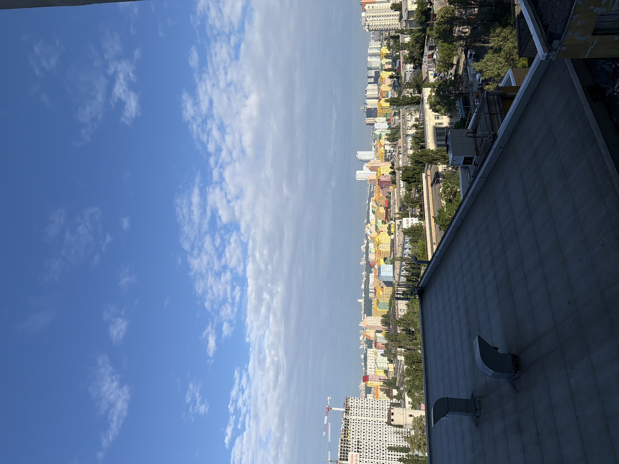 The image is an elevated view of a city skyline, likely Kyiv, Ukraine, dominated by a bright blue sky with scattered white clouds. In the foreground, a dark gray, flat roof occupies the bottom third of the frame, featuring two gray ventilation pipes. Beyond the roof, the middle ground displays a dense urban landscape with buildings of varying heights and architectural styles.
A prominent feature is a construction site on the left, with a tall white building under construction and two construction cranes. Adjacent to it, a cluster of brightly colored apartment buildings, painted in shades of yellow, pink, blue, and green, forms a vibrant section of the cityscape. These colorful buildings suggest a residential area with modern architecture.
Further in the distance, a wider panorama of the city unfolds, with more buildings stretching towards the horizon. Trees, primarily deciduous and with green foliage, are interspersed throughout the urban fabric, indicating a presence of parks or tree-lined streets. The overall impression is of a bustling city on a clear day, with a mix of older and newer construction. The angle suggests the photograph was taken from a high vantage point, possibly from another building.