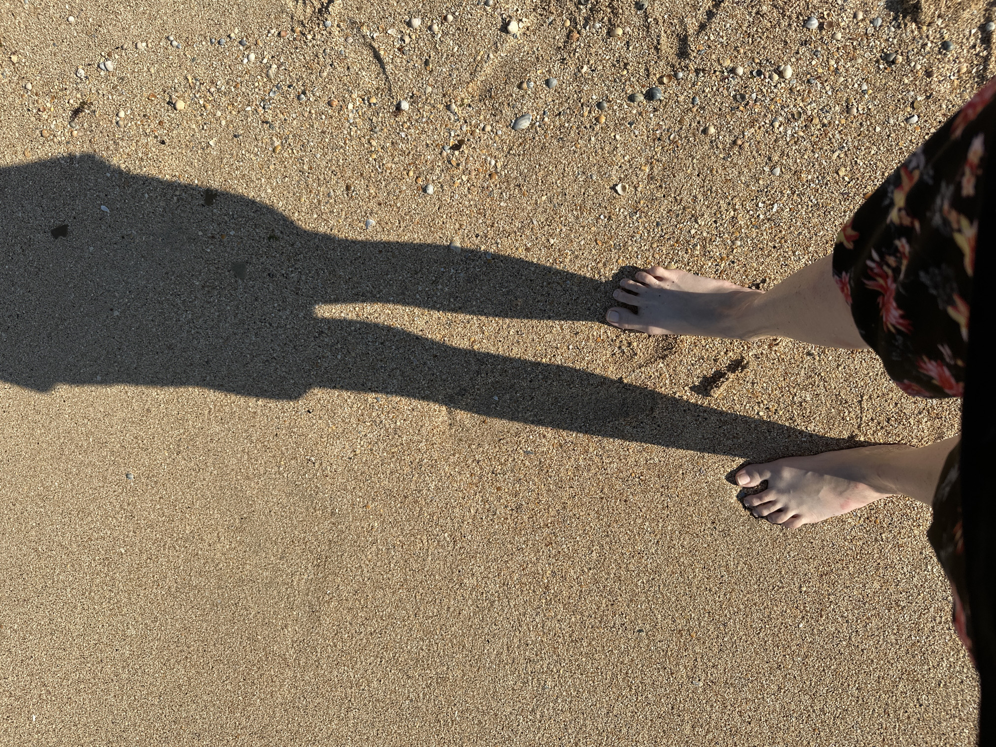 This media file features a person's bare feet on a sandy beach. The sand is light brown and speckled with small pebbles and shell fragments. A long shadow of the person stretches across the sand from the left side of the frame, indicating the sun is casting a distinct shadow. The person's legs are visible from the knees down, with one foot closer to the camera on the right, and the other further away on the left, aligned with the shadow. The person is wearing a dark garment with a floral pattern. The scene suggests a warm, sunny day, likely during daylight hours, given the strong shadow. The context of Golubitskaya, Russia, places this scene on a beach. No text is visible.