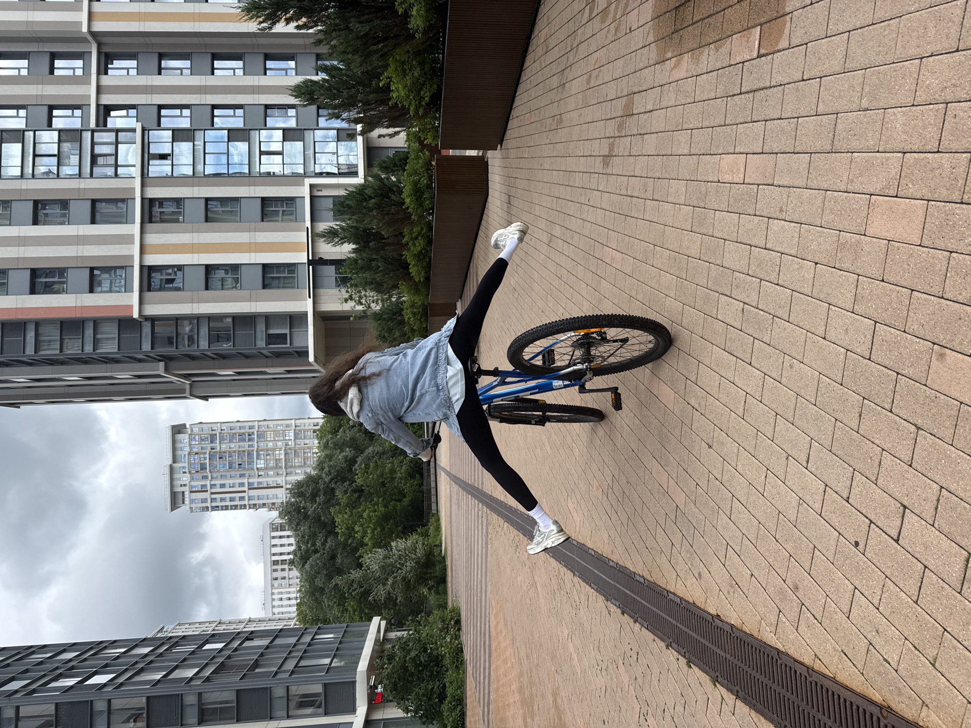 A person is seen from behind riding a blue bicycle, with their legs spread wide on either side of the bike. They are wearing a denim jacket, a light-colored shirt, black leggings, white socks, and white sneakers. The bicycle has black tires and a silver frame. The person is in motion, with their right leg extended forward and their left leg extended backward.
The scene appears to be an outdoor urban environment, likely a pedestrian walkway or a plaza. The ground is paved with light-colored rectangular bricks arranged in a pattern. A metal drainage grate runs along the left side of the walkway.
In the background, there are modern, tall residential buildings with distinctive facades featuring vertical bands of various colors, including grey, beige, and orange. The buildings have numerous windows that reflect the cloudy sky. Rows of dark green evergreen trees and shrubs are planted in raised planters between the walkway and the buildings. The sky is overcast with gray clouds. The overall impression is a contemporary urban setting.
The image does not provide any specific information or visual cues that would allow for a definitive identification of the location as Moscow, Russia.