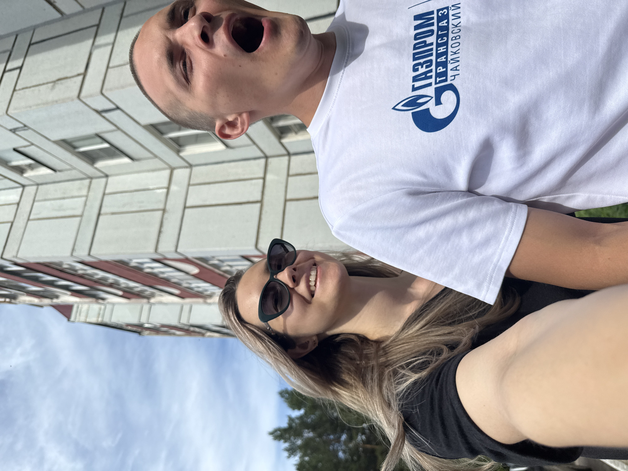 The image features two people outdoors under a bright sky, appearing to take a selfie or be photographed together from a low angle. The photo is oriented vertically, with the subjects appearing rotated on their sides.
The person on the left (when rotated to upright view) is a woman with long, light-colored hair, wearing black sunglasses and a black top. She is smiling broadly, revealing her teeth, and appears happy. Her arm, likely holding the camera, is visible in the foreground.
The person on the right (when rotated upright) is a man with very short, buzzed hair. He is wearing a white t-shirt with a blue logo and text. His mouth is wide open in a dramatic, perhaps playful or surprised expression, and his eyes are also open wide. The text on his t-shirt reads "ГАЗПРОМ ТРАНСГАЗ ЧАЙКОВСКИЙ" (Gazprom Transgaz Chaikovsky), indicating a connection to the city of Chaikovsky, Russia, and the company Gazprom.
In the background, a multi-story building with a light-colored facade and visible window frames can be seen, suggesting an urban or residential setting. The sky is bright blue with scattered white clouds, indicating clear and sunny weather. Some blurry green foliage, possibly trees, are visible in the distance. The strong sunlight casts shadows, suggesting it is daytime. The overall mood appears casual and joyful.