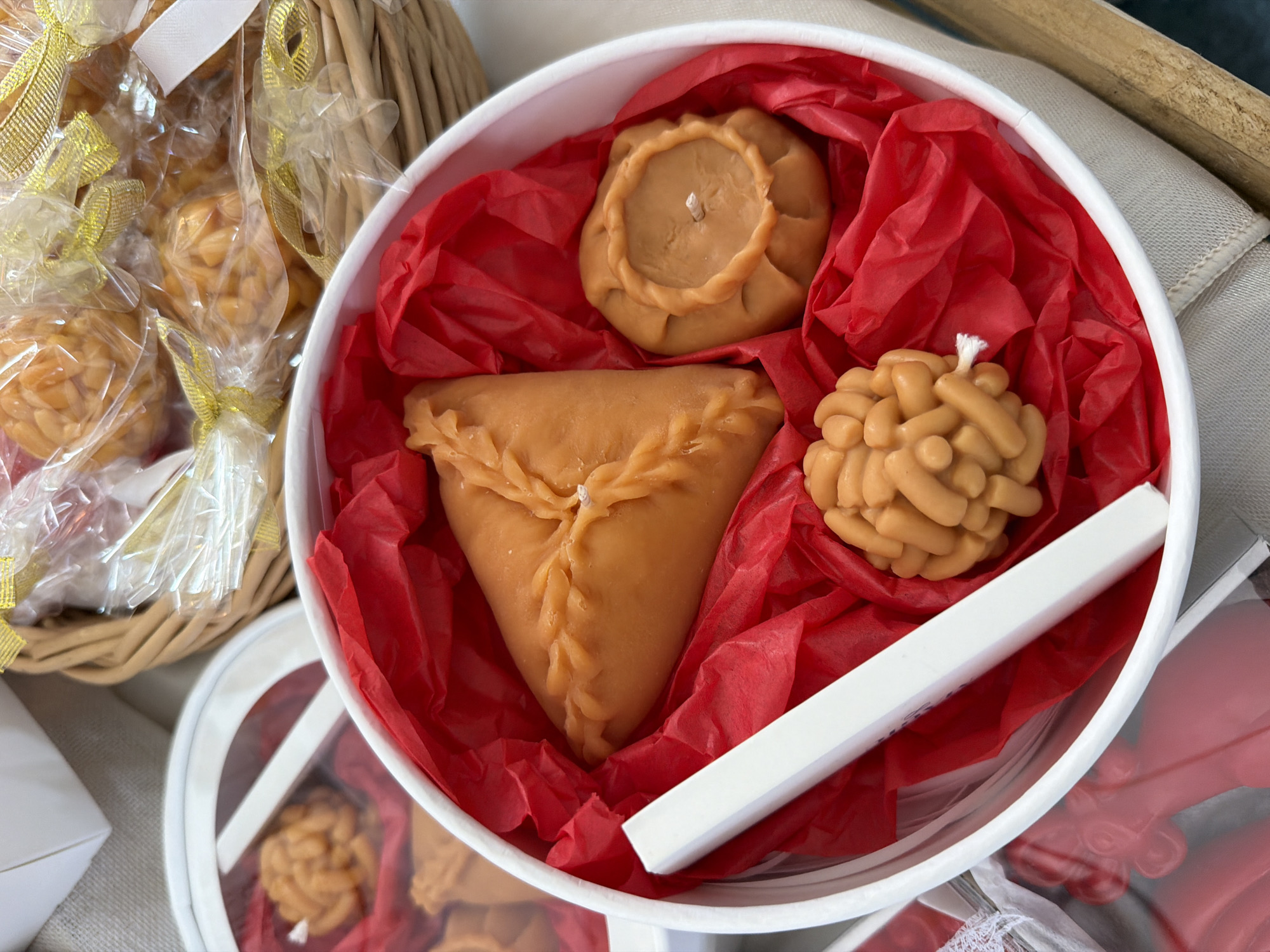 In an overhead shot, a close-up view showcases a white cylindrical container filled with red tissue paper, holding three intricately shaped beeswax candles that resemble pastries. One candle, triangular in shape with a crimped edge, is positioned on the left. To its right sits a round candle with a decorative rim. At the bottom of the container is a spherical candle with a textured, segmented surface. A white rectangular object, possibly a wick or a decorative element, lies diagonally across the container, in front of the triangular candle. In the upper right quadrant of the image, a woven basket filled with similarly shaped, individually wrapped candles tied with gold ribbon adds depth to the composition. Another white container, partially visible on the left, also contains what appear to be more of these pastry-like candles. The background is a neutral, textured surface.