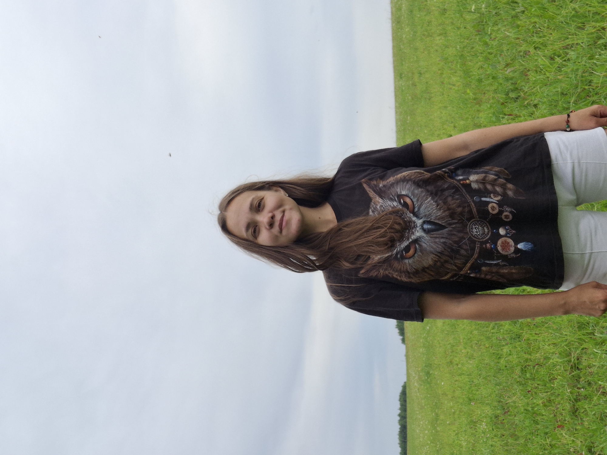 A young woman with long brown hair stands in a grassy field under a cloudy sky. She is wearing a black t-shirt with a large owl graphic on the front and white shorts. A bracelet is visible on her left wrist. The field is green and appears to be a rural area. The horizon line is visible in the distance with a line of trees. The image does not provide any specific information to suggest the location is the city of Гаревая, Russia.