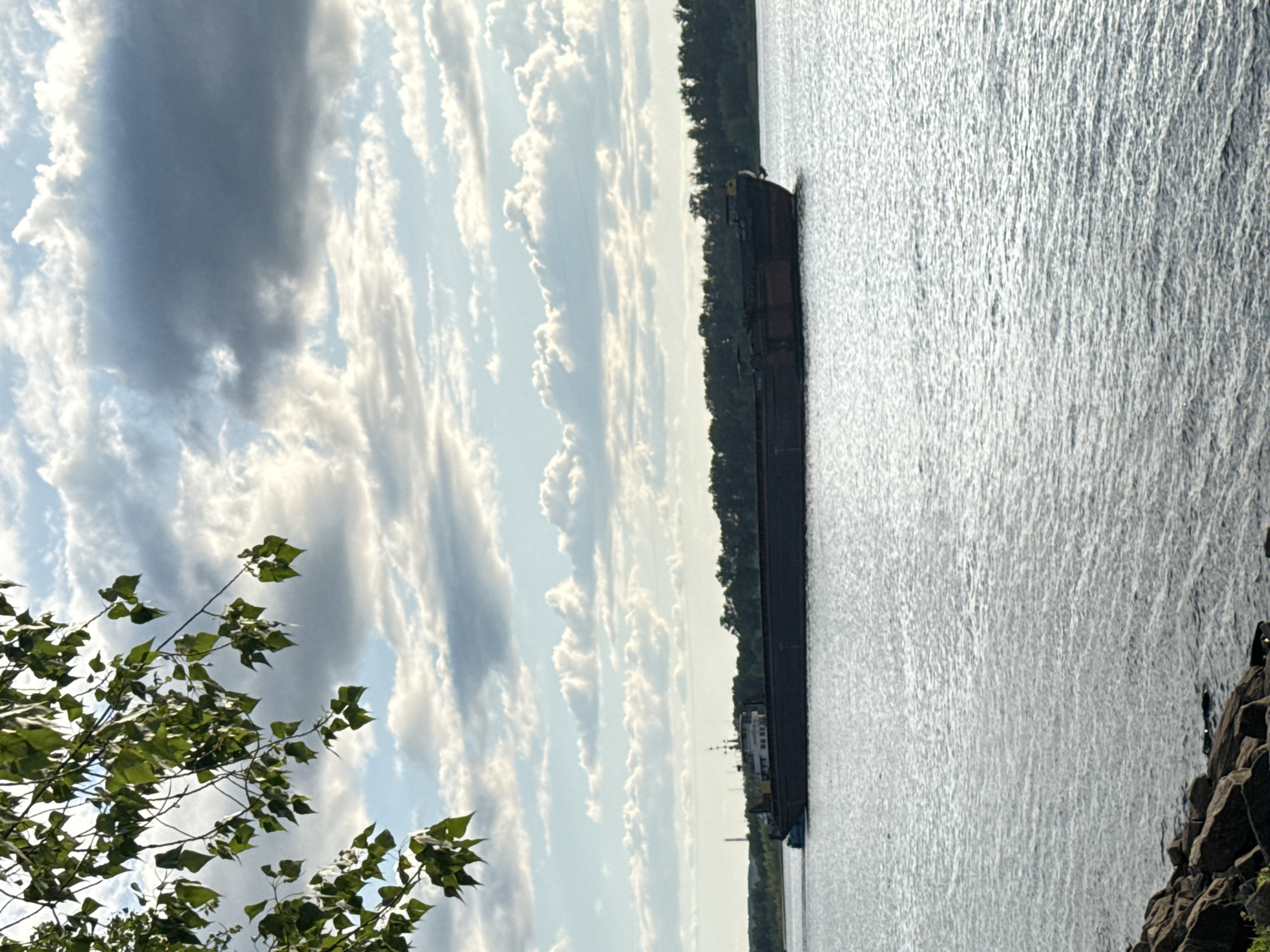 The image depicts a serene river scene on a cloudy day. In the foreground, a rocky shoreline with some green vegetation is visible. The middle ground is dominated by the calm, rippling surface of a wide river. A long, dark barge with a small cabin structure at the stern is moving across the water, heading from left to right. In the background, a dense line of green trees marks the opposite bank of the river, and the sky is filled with a mix of bright white and dark grey clouds. The overall impression is one of natural beauty and quietude, with human activity represented by the passing barge. The location is identified as the Kama River in Chaikovsky, Russia, a city known for its industrial significance and its position on the river.