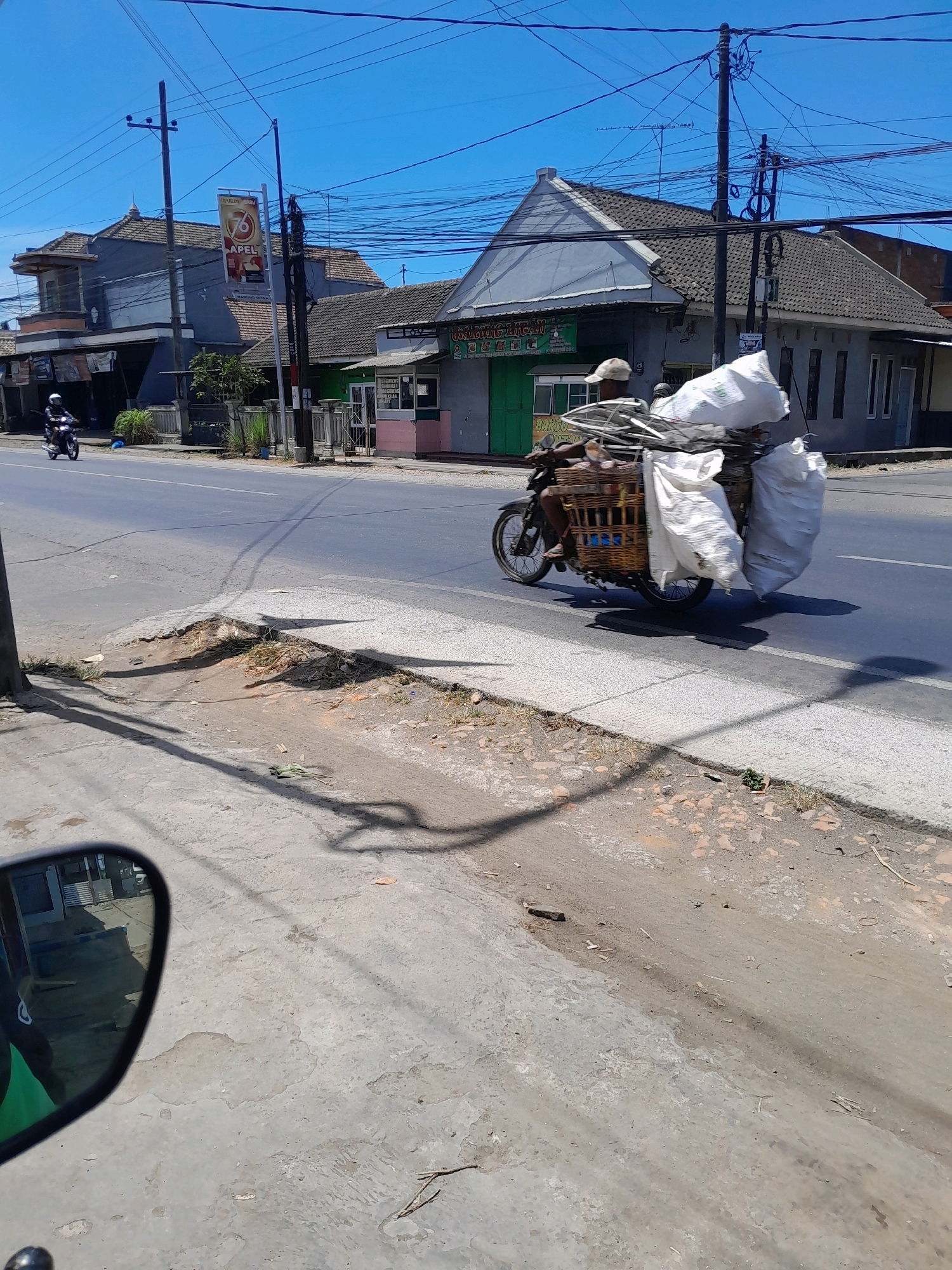 In Talangagung, Indonesia, a motorcycle carries a substantial load of recycled materials. The rider, wearing a cap, is seated on the motorcycle, which is equipped with a large woven basket and several large white bags strapped to the back. These bags appear to be filled with a mix of metal scraps and possibly other discarded items. The motorcycle is traveling on a paved road, with buildings visible on the side of the road, including what appears to be a business with green doors and signs. The sky is clear and blue, indicating a sunny day. The foreground shows a concrete sidewalk and a patch of dirt with some vegetation, with shadows cast across it from overhead power lines and the motorcycle. The overall scene depicts everyday life and commerce in a rural or semi-urban Indonesian setting.
