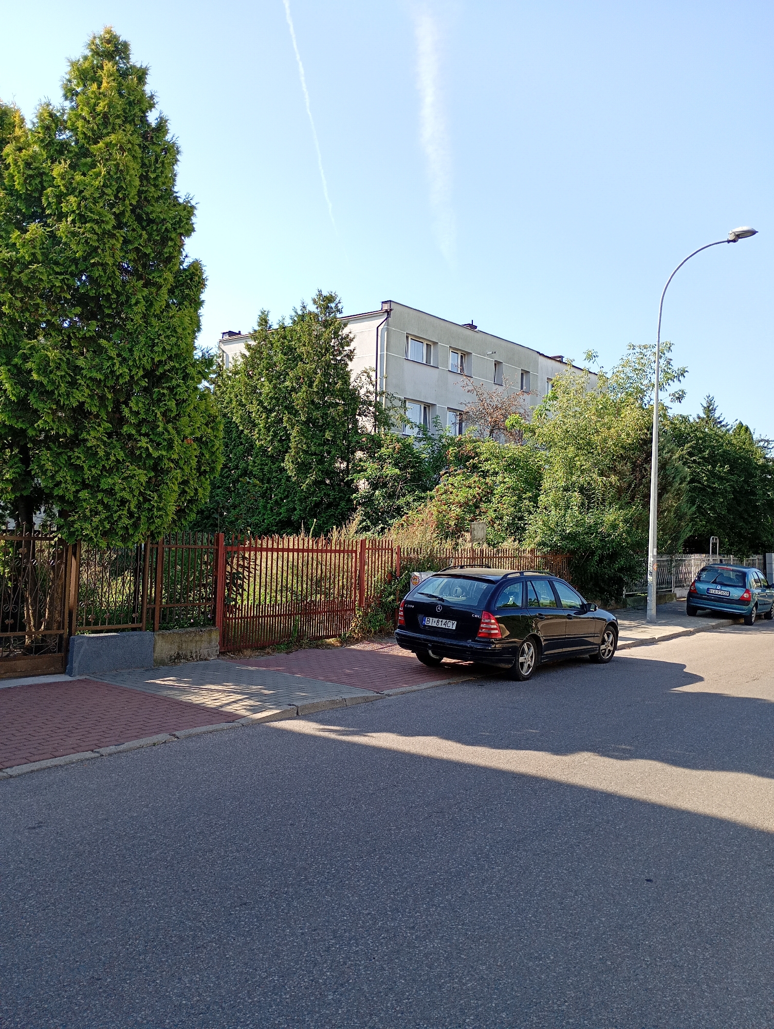 This image shows a street scene in Białystok, Poland, on a clear day. In the foreground, a dark blue Mercedes-Benz station wagon is parked on the right side of the asphalt road. The car has a license plate that reads "BI 814CY". Further down the road, a smaller, teal-colored hatchback is also parked, with its license plate visible as "BIA 95659".
Behind the parked cars, a pedestrian walkway made of red and gray bricks runs alongside a rusted metal fence. The fence encloses a property with abundant green foliage and trees, including a tall, conical evergreen tree on the left. Beyond the trees, a multi-story, light gray apartment building with numerous windows is visible. A street lamp with a curved pole stands on the right side of the walkway, casting a shadow on the road. The sky is a clear, bright blue with a faint white contrail from an airplane.