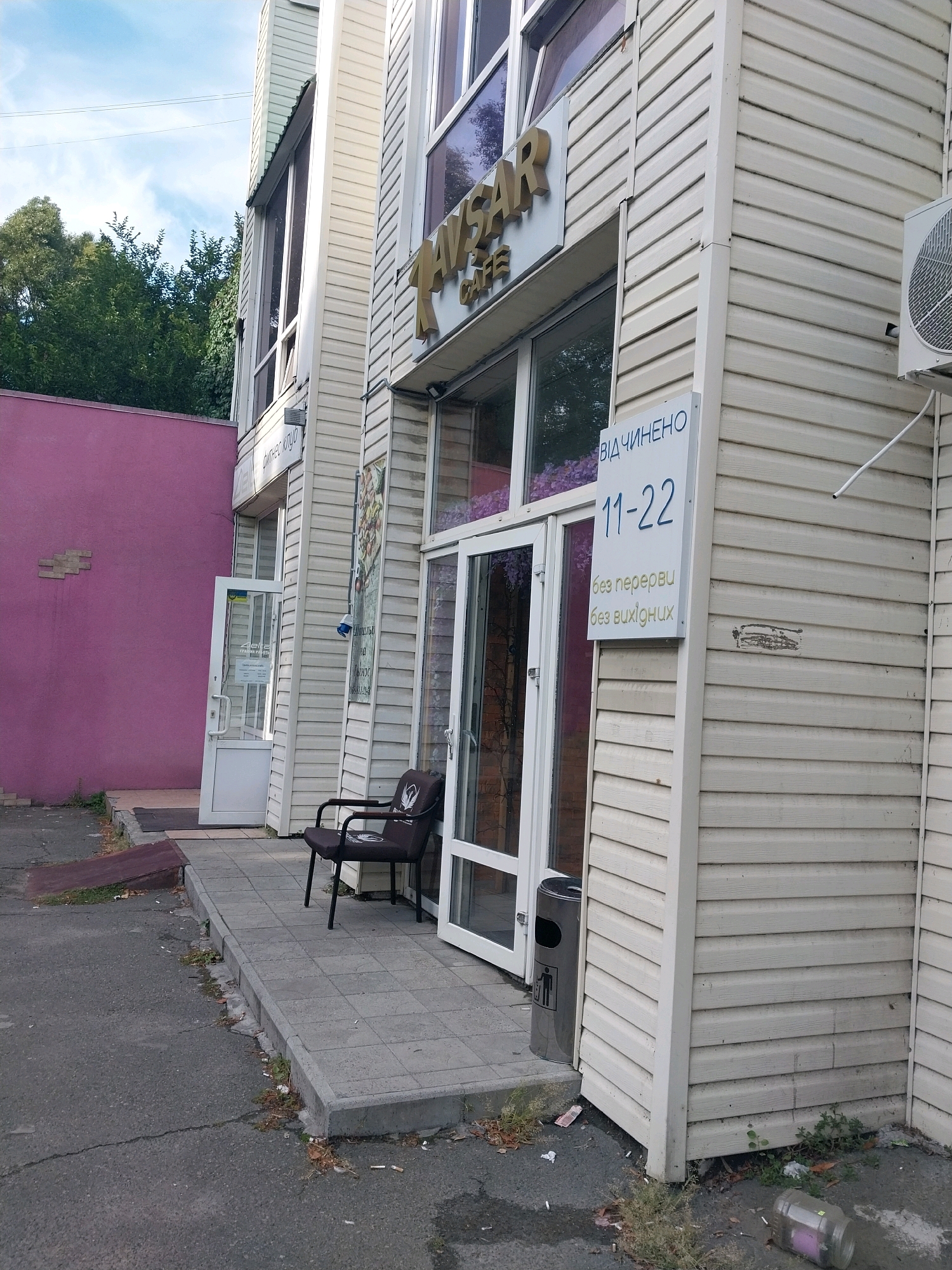 The image shows the exterior of a cafe named "TAVSAR CAFE" located on the first floor of a building. The building is clad in light-colored horizontal siding. To the left of the cafe entrance, there is a pink wall. The cafe has large windows and glass doors. A sign on the building indicates operating hours: "11-22" and "без перерви без вихідних" (without breaks, without days off). A dark chair with a logo is placed on the tiled walkway in front of the cafe. A metal trash can is located near the entrance. The scene appears to be in a commercial area, with a paved area in front of the building and some greenery. The overall condition of the area suggests it is an outdoor public space.