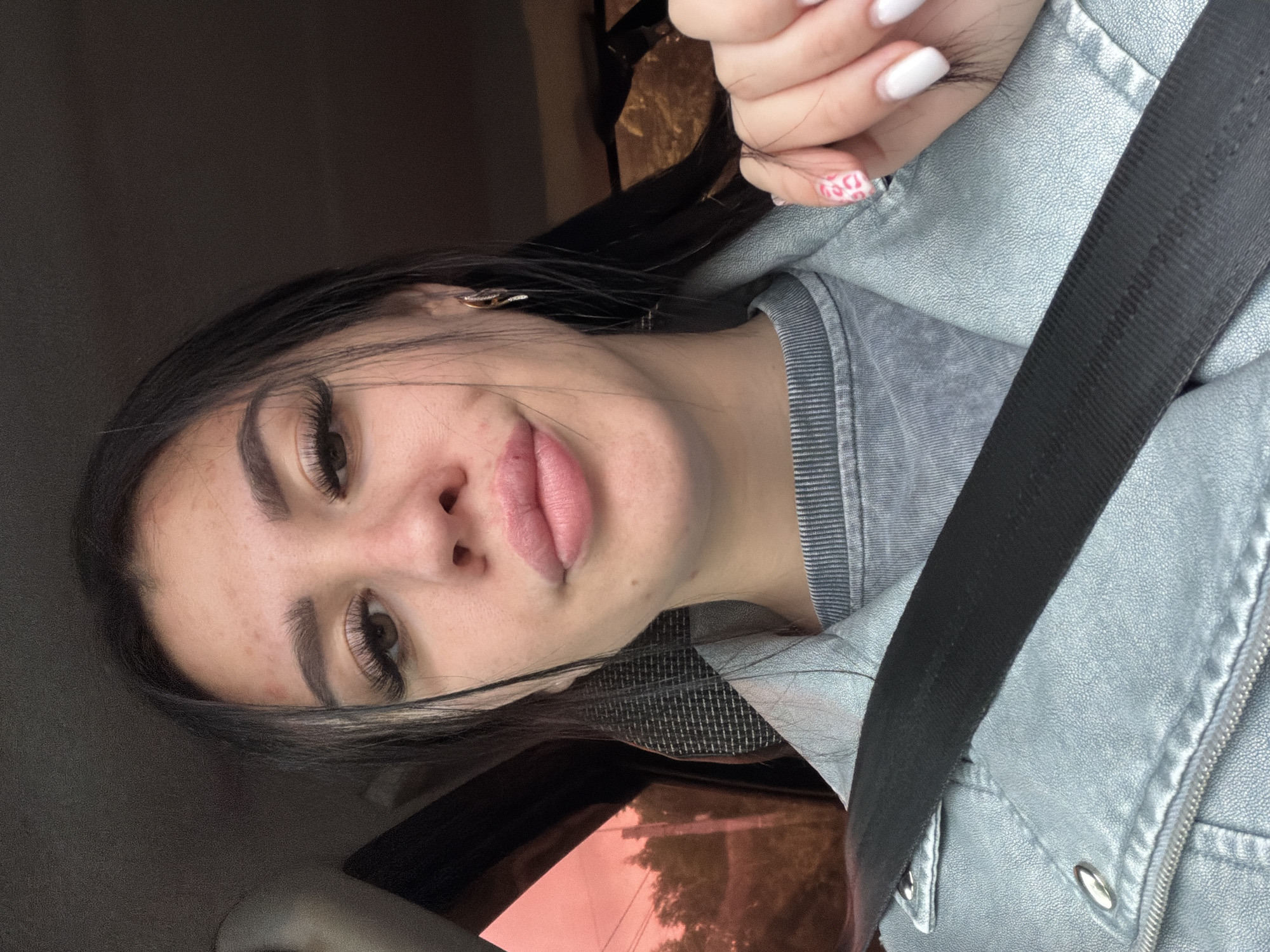 The scene is set within a vehicle, featuring a young adult female, in the city of Chaikovsky, Russia. The woman is photographed in a close-up, chest-up view, looking directly at the camera. She has dark hair, styled with some strands falling over her face, and visible brown eyes with long, dark eyelashes. Her eyebrows are well-defined. Her lips are full, appearing slightly pouted, with a pink-toned lipstick. Minor blemishes are present on her forehead and cheeks. She wears a light blue-grey textured jacket over a grey ribbed crew-neck t-shirt. A black safety belt crosses diagonally from her right shoulder across her chest. She wears small gold hoop earrings with white stones. Her right hand, with white nail polish on most fingers and a pink and white patterned nail on the ring finger, is visible near her right shoulder. Through the vehicle's left-side window, an exterior view tinted reddish-orange shows the indistinct outlines of trees and potentially utility poles against a lighter sky. The vehicle's interior appears dark.