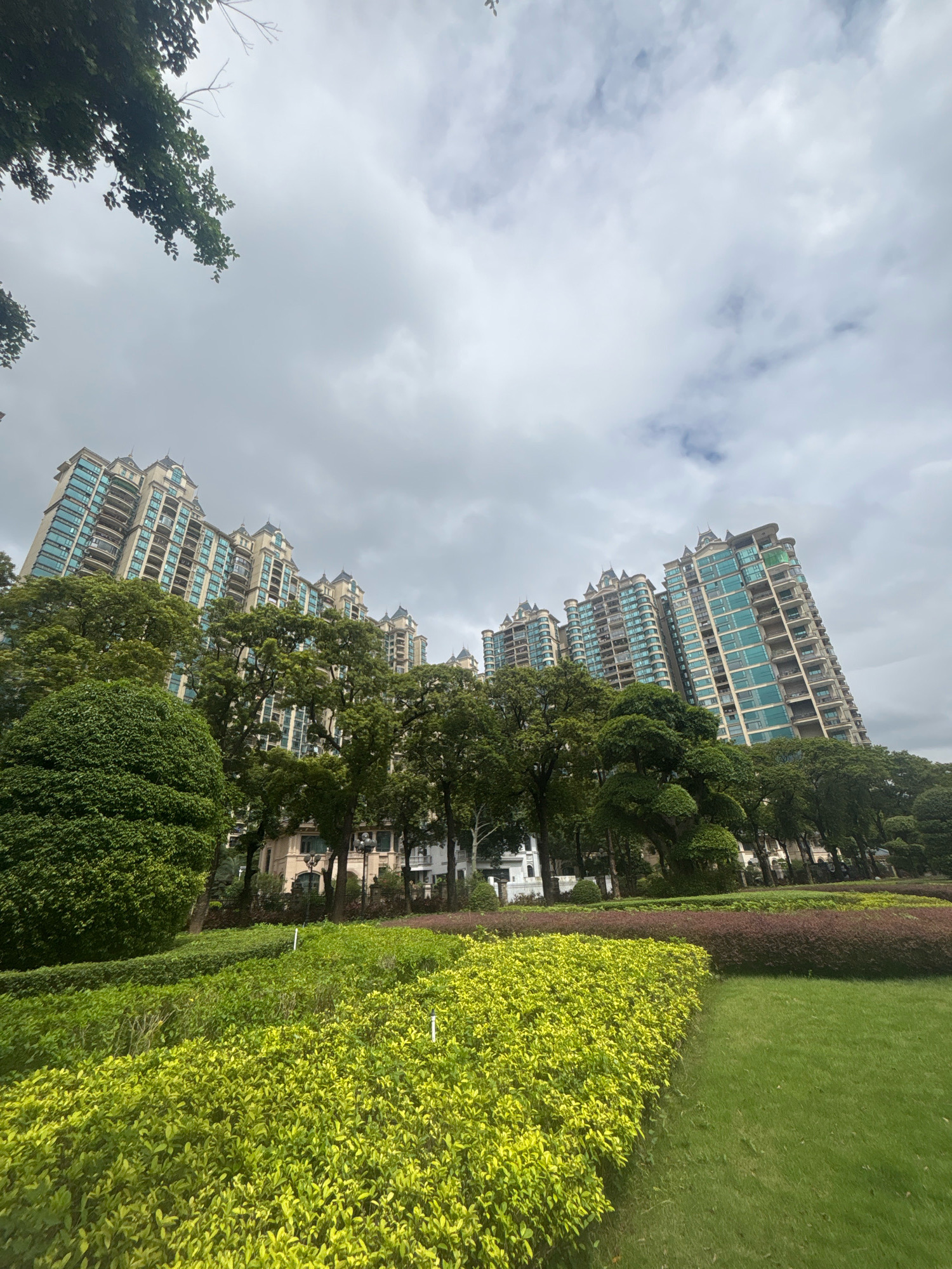 This is an outdoor, low-angle, wide shot of apartment buildings and landscaping in Nanhai District, China. The scene is dominated by a row of tall, multi-story apartment buildings with light-colored facades and turquoise glass windows. They have ornate rooflines with decorative spires. In front of the buildings is a dense canopy of lush green trees. Below the trees, meticulously manicured hedges and grassy areas form a park-like setting. The sky above is overcast with heavy gray clouds, suggesting a cloudy or possibly rainy day. There are no people visible in the image. The overall impression is one of a well-maintained urban residential area.