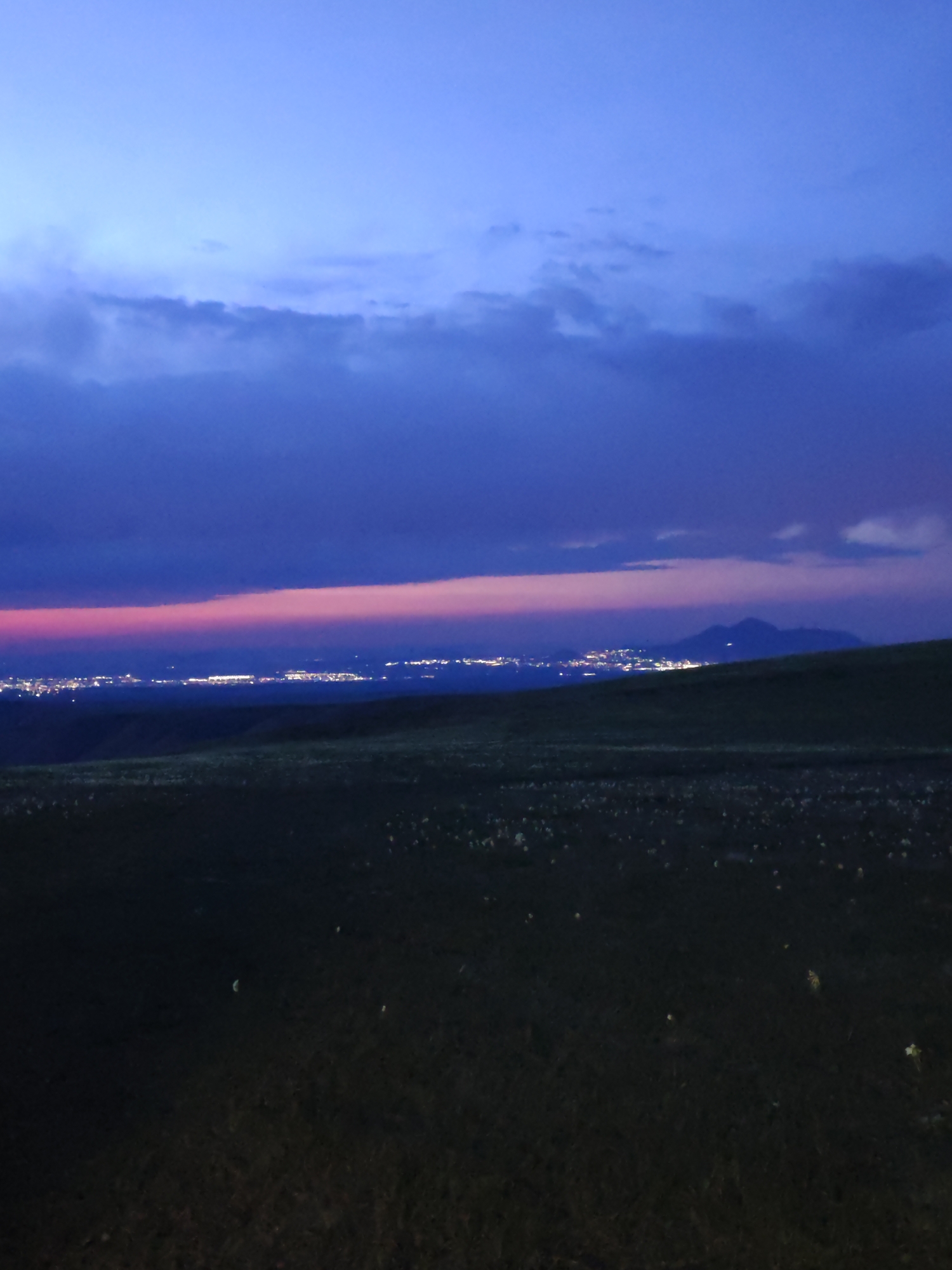 The image shows a landscape at dusk with a dark field in the foreground. In the distance, a city is visible with its lights illuminating the horizon. Above the city, a band of pink and purple hues is present in the sky, transitioning into darker blue clouds. The sky is mostly covered with dark blue clouds. A hill is visible on the right side of the image.