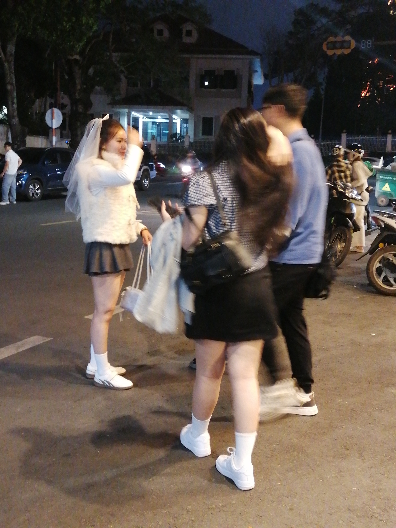 The image captures a night scene on a street in Da Lat, Vietnam, likely near the city center given the presence of a traffic light with the number "88" displayed. In the foreground, a woman wearing a white veil, a fluffy white vest over a long-sleeved white top, and a short gray pleated skirt stands with her legs slightly apart. She has white socks and white shoes. She is holding a light-colored bag in her right hand. To her left, a blue car is parked, and a person in a white shirt and blue pants is partially visible.
Behind the woman in the veil, another woman is visible from the side, wearing a black and white checkered shirt and a short black skirt. She is also wearing white socks and white shoes. To the right of these two women, a man wearing a blue long-sleeved shirt and black pants stands with his right arm slightly raised. His left hand is obscured. Further in the background, several motorcycles and people are visible, some with helmets. A green cart or vehicle is also present to the right. The street is paved, and shadows are cast on the ground. The atmosphere is dimly lit, suggesting nighttime, with streetlights and building lights providing illumination. The architecture in the background suggests a residential or commercial building.
