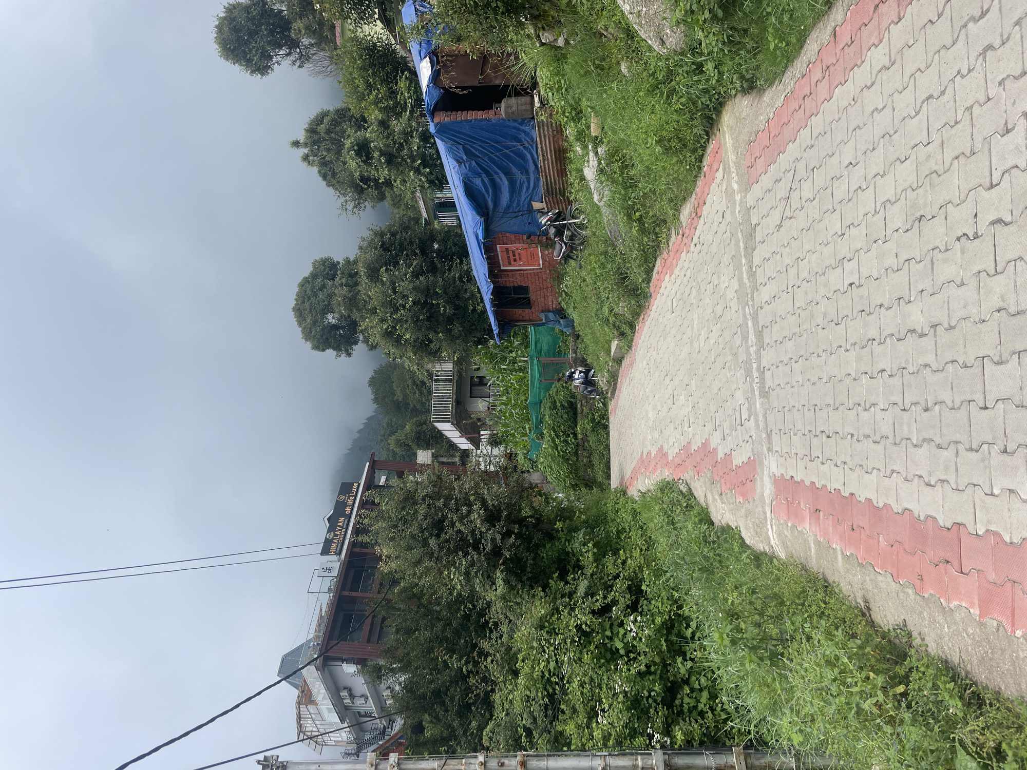 This image depicts a paved pathway in Dharamkot, India, curving uphill and lined with greenery. A multi-story building with "HIMALAYAN CROWN" signage is visible on the left, surrounded by trees. To the right, a small brick structure is covered by a blue tarp, with a motorcycle parked nearby. The sky is overcast, contributing to a muted atmosphere. The pathway is constructed from interlocking pavers with a red border. In the middle distance, two individuals are seated on the side of the path.