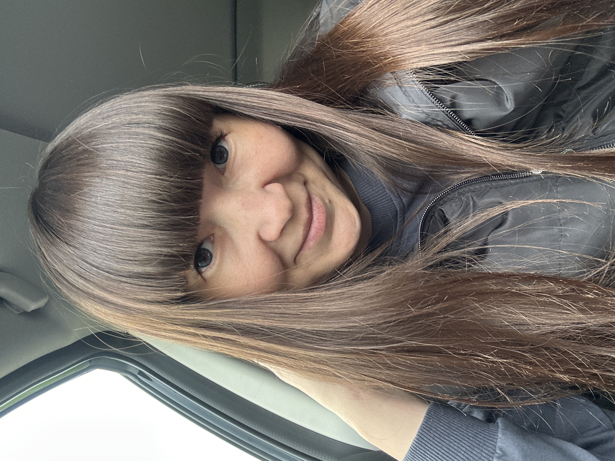 A woman with long, straight brown hair and bangs is seated in a vehicle. She is wearing a black jacket over a gray sweatshirt. The woman is smiling slightly at the camera. The interior of the vehicle is visible, including the window and dashboard. The location is not discernible from the image. No information regarding Kamennoye, Russia is discernible from the image.