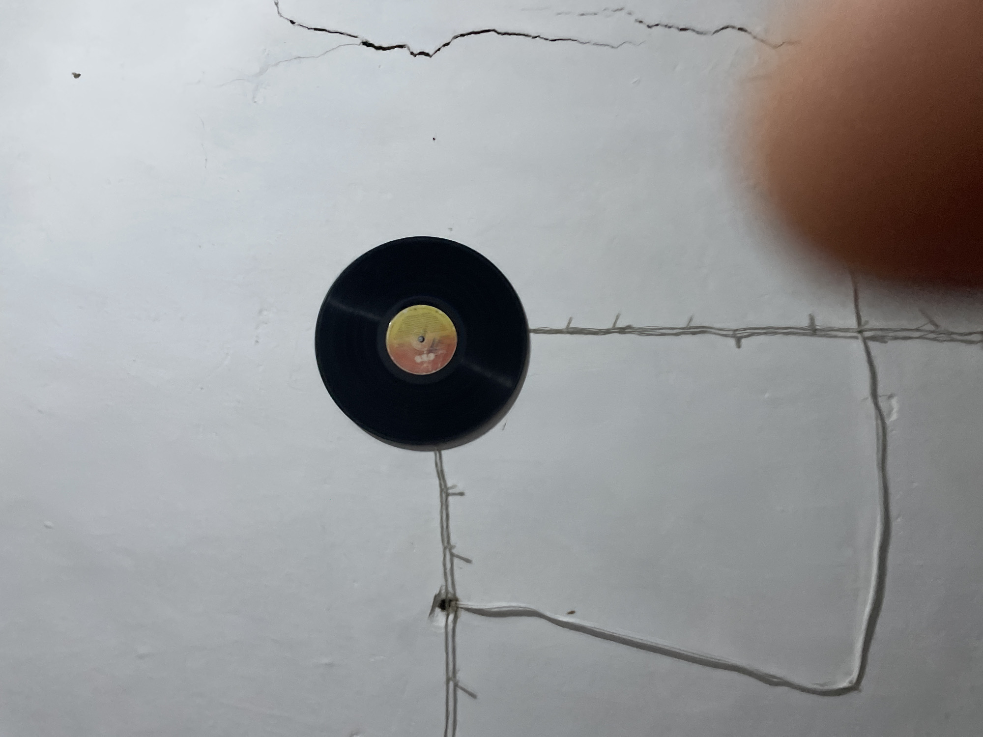 A vinyl record is suspended from a string of lights against a textured, white wall. The record is black with a yellow and orange label in the center. The string of lights is also white and is draped across the wall, attached to the wall with small clips. There are several cracks in the wall, one of which is quite prominent and runs vertically on the right side of the image. In the lower right corner, a blurry, reddish-brown object, likely a person's finger, obstructs a portion of the scene. The background is a plain, white wall with a rough texture.