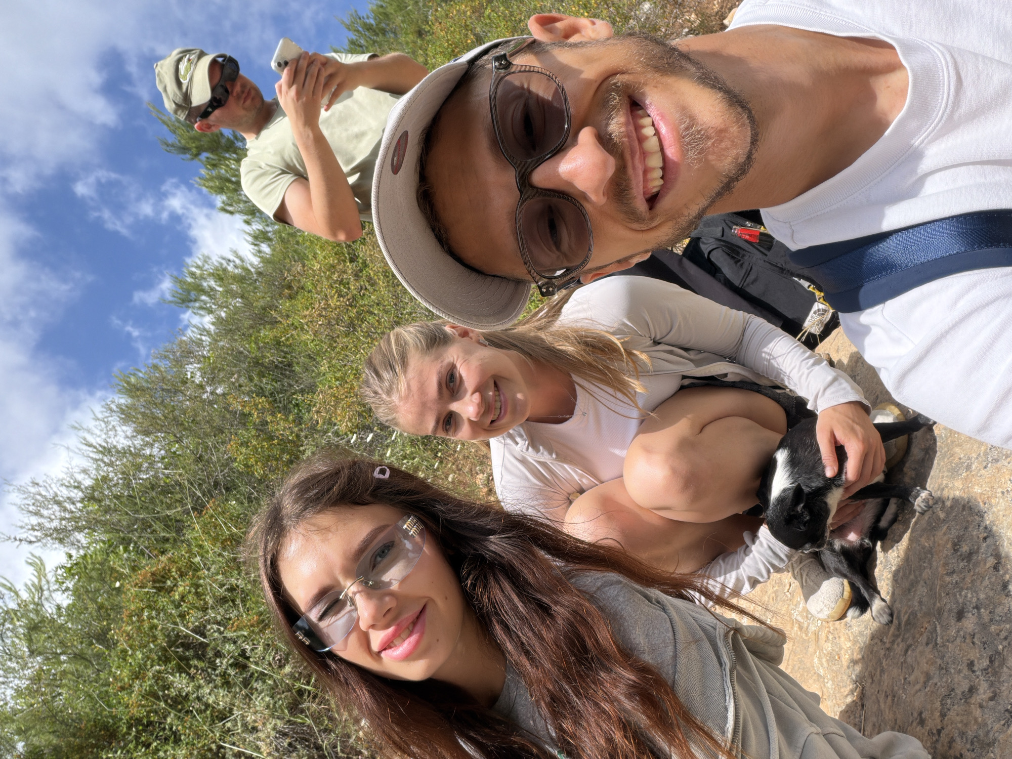 A medium shot shows a group of four people taking a selfie with a black and white dog outdoors in Tirat Karmel, Israel.
A man with short brown hair and a beard wears a white hat, sunglasses, and a white t-shirt in the upper right. The selfie is taken from his perspective. To the left and slightly below him is a woman with blonde hair wearing a white t-shirt and a light jacket. She is crouched down and holding the dog. In the lower left is a woman with long brown hair wearing glasses and a grey sweatshirt. Behind the group is a man wearing a green hat, sunglasses, and a green t-shirt, looking at his phone.
The background consists of green bushes and trees, a blue sky with white clouds, and rocky ground.