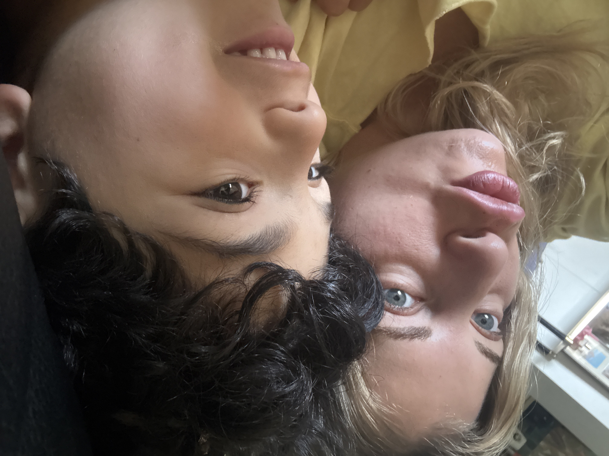 A selfie-style photograph shows two people lying head-to-head. On the left is a child with dark, curly hair, brown eyes, and tan skin. They are smiling slightly, revealing their teeth. On the right is an adult with blonde hair, blue eyes, and fair skin. Both appear to be wearing yellow clothing. The background is blurred, but a picture frame and furniture are visible, which is located in Rousset, France.