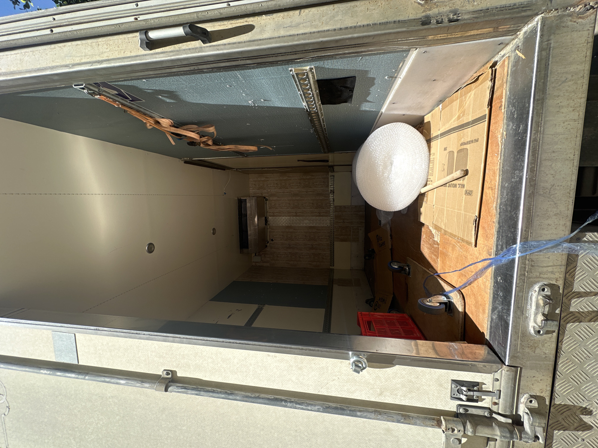 The image shows the interior of an empty truck box, likely a refrigerated truck, viewed from the open rear doors. The truck is parked outdoors, with sunlight casting shadows inside. The interior walls are a light beige on the left and a blueish-gray on the right. There are some straps hanging from the right wall and a metal track system along the same wall. The floor of the truck is made of wood and appears to be covered in cardboard boxes. A red plastic crate is visible on the left side of the floor, near the rear door. A roll of bubble wrap and a wooden dowel are placed on a cardboard box in the center of the floor. There are two casters visible on the floor, one attached to a piece of cardboard and another under the red crate. The truck appears to be in a state of being emptied or prepared for transport. The location is not identifiable from the image itself, and no specific details about Tel-Aviv, Israel can be inferred.