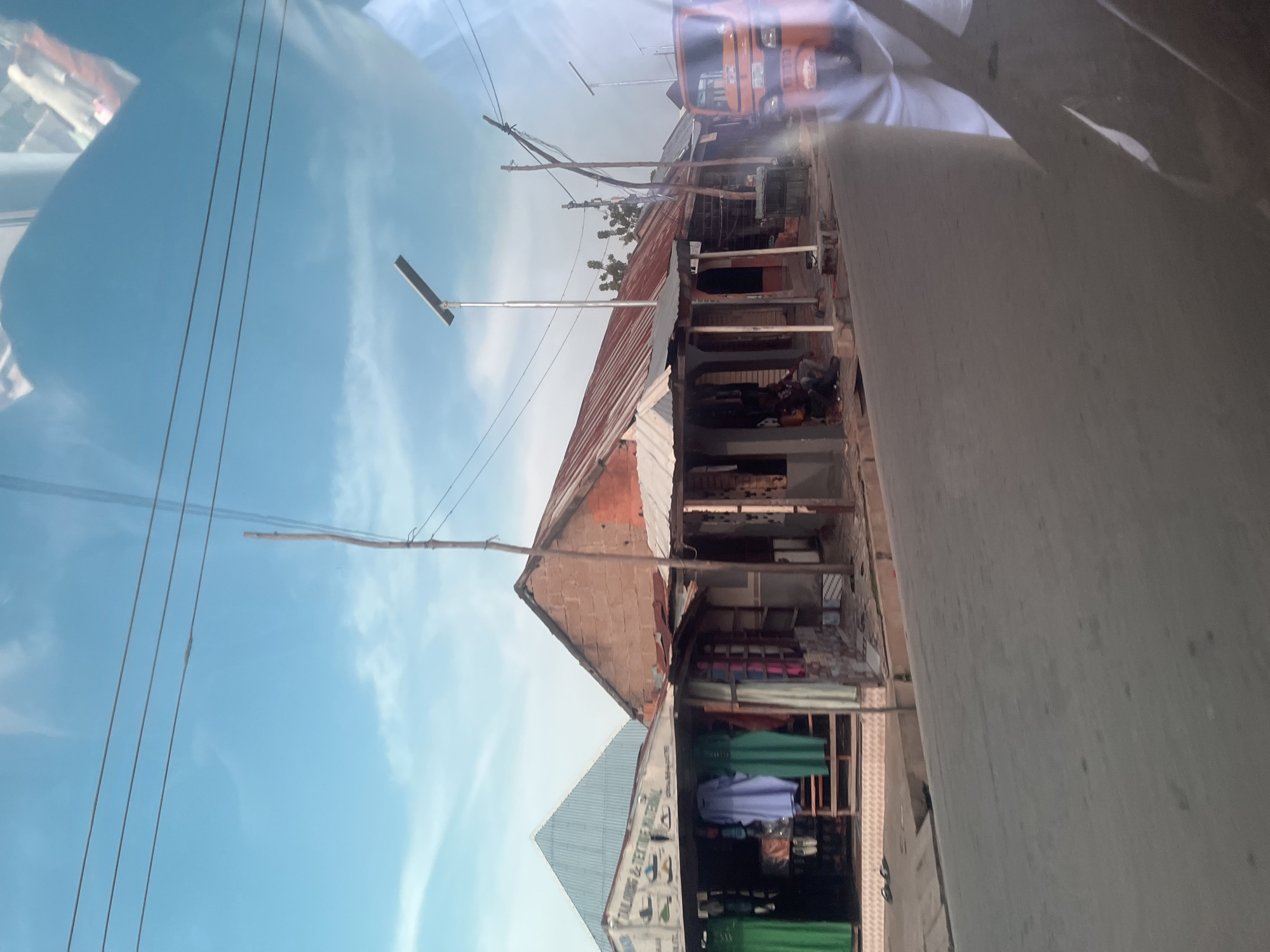 This media file captures an outdoor scene in Jalingo, Nigeria. The primary subjects are a row of shops with corrugated metal roofs, some of which appear to be made of brick. The shops are situated along a paved road, suggesting a commercial area. In front of the shops, colorful textiles such as green and purple fabrics are displayed, indicating that these are likely clothing or fabric stores. Some of the shops have open fronts, revealing shelves stocked with various items.
Visible objects include a solar-powered street light with a rectangular panel, several utility poles with wires, and what appears to be an orange auto-rickshaw (a tuk-tuk) parked nearby. The reflection on the surface in the foreground, possibly a vehicle's window or a protective covering, shows a blurred image of people wearing white clothing.
The sky is bright blue with scattered white clouds, suggesting it is daytime with fair weather. The scene depicts a typical street view of a commercial district in Nigeria, with the buildings showcasing a rustic and somewhat worn appearance. There is some visible text on a sign above one of the shops, which includes the words "TAILORS & TEXTILE MATERIAL". The overall impression is one of a busy, everyday commercial environment.