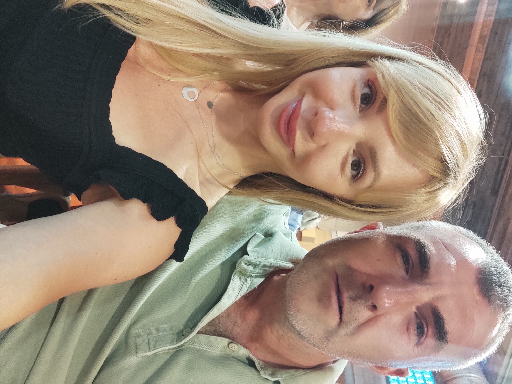 The image is a selfie taken by a man and a woman, with another woman blurred in the background. The man, on the right, has short gray hair and is wearing a light green polo shirt. The woman in the foreground, on the left, has long blonde hair and is wearing a black top. They appear to be sitting at a table in a restaurant, with wooden beams visible on the ceiling, suggesting an indoor setting. The woman in the background is out of focus. The image does not contain enough information to determine the specific location within Blanes, Spain.